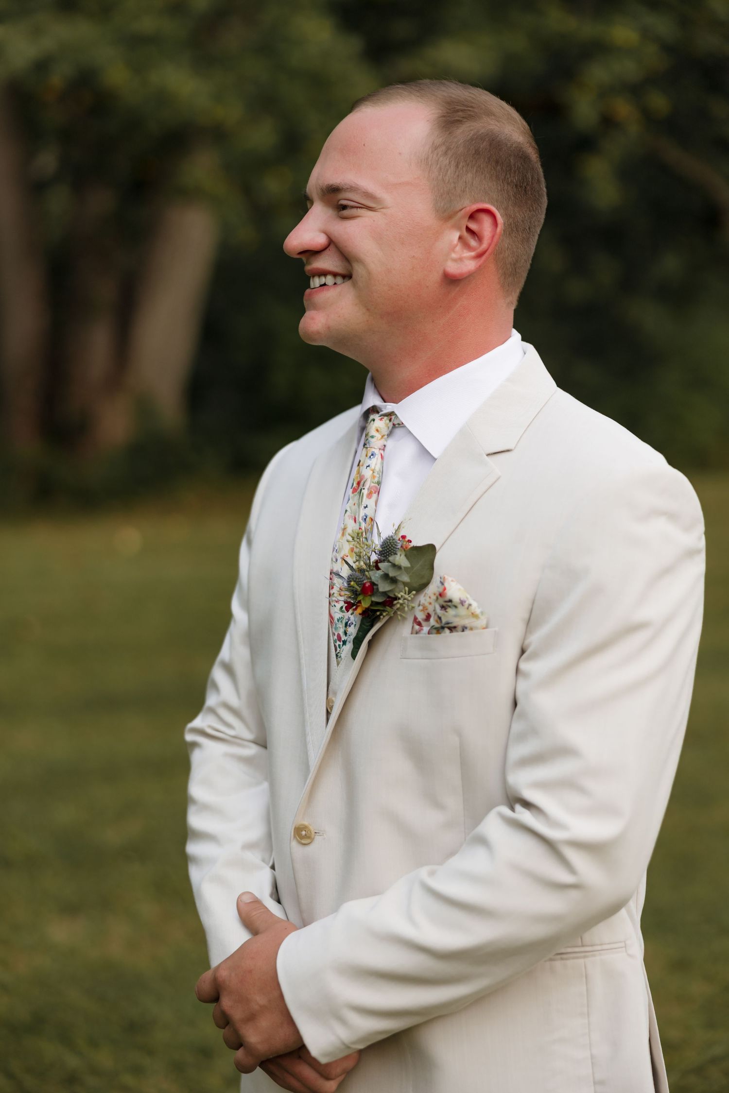 Smiling groom in ivory suit with floral tie and boutonniere standing outdoors at a wedding ceremony.