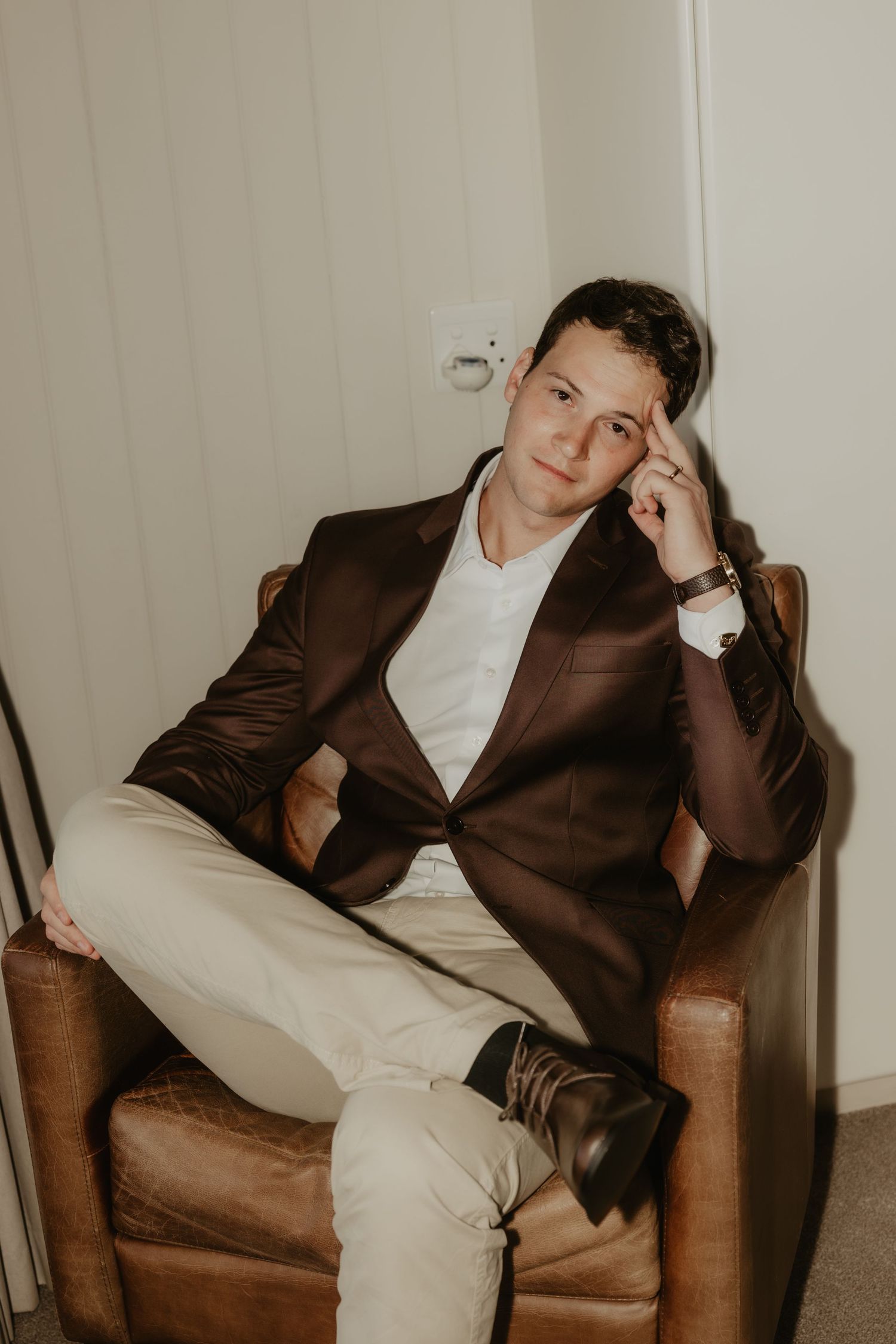 Young man in brown blazer and white shirt sits thoughtfully in leather chair, resting hand on temple.