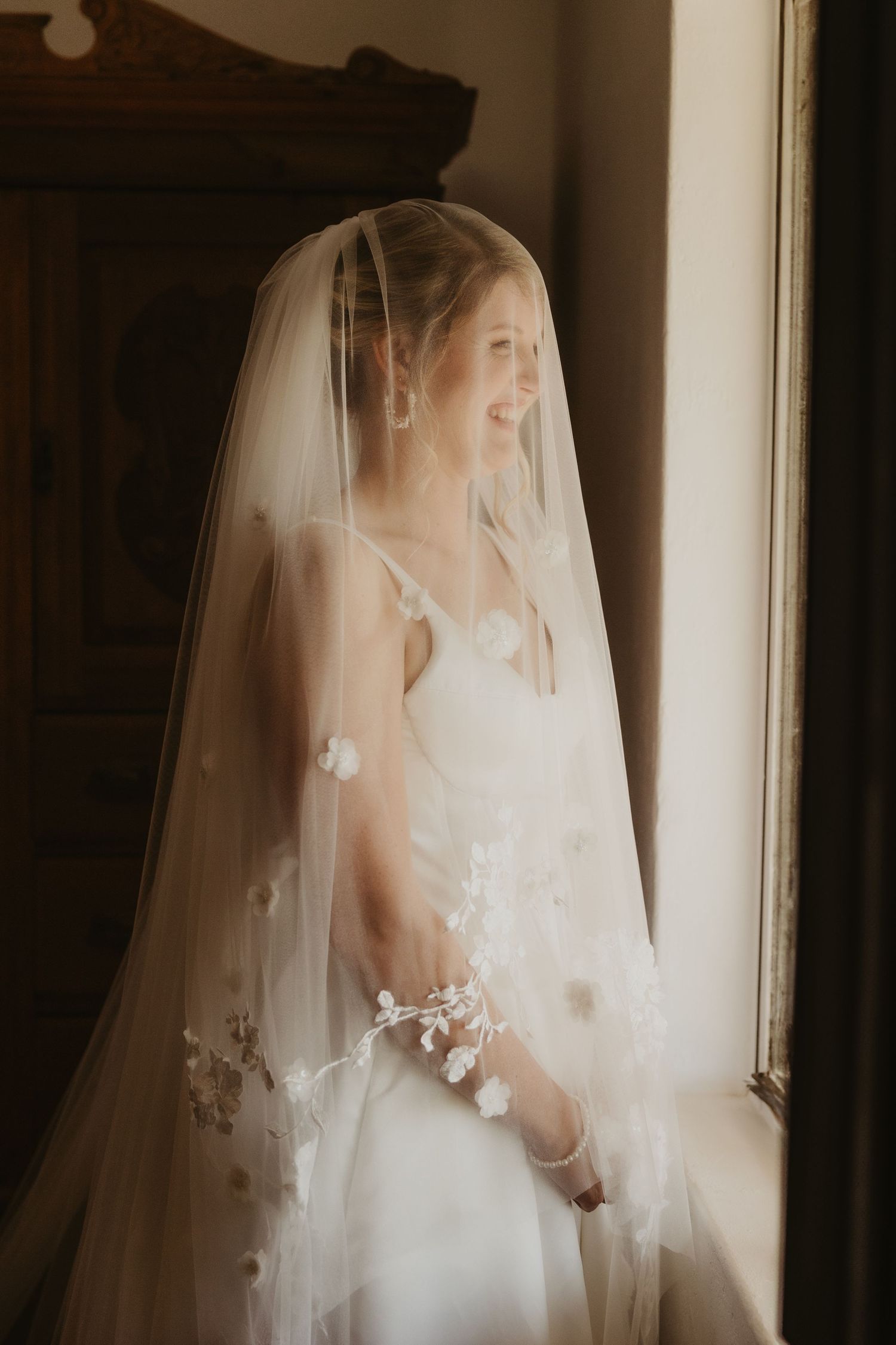 Smiling bride in white wedding dress and floral veil standing by a sunlit window in soft natural light.