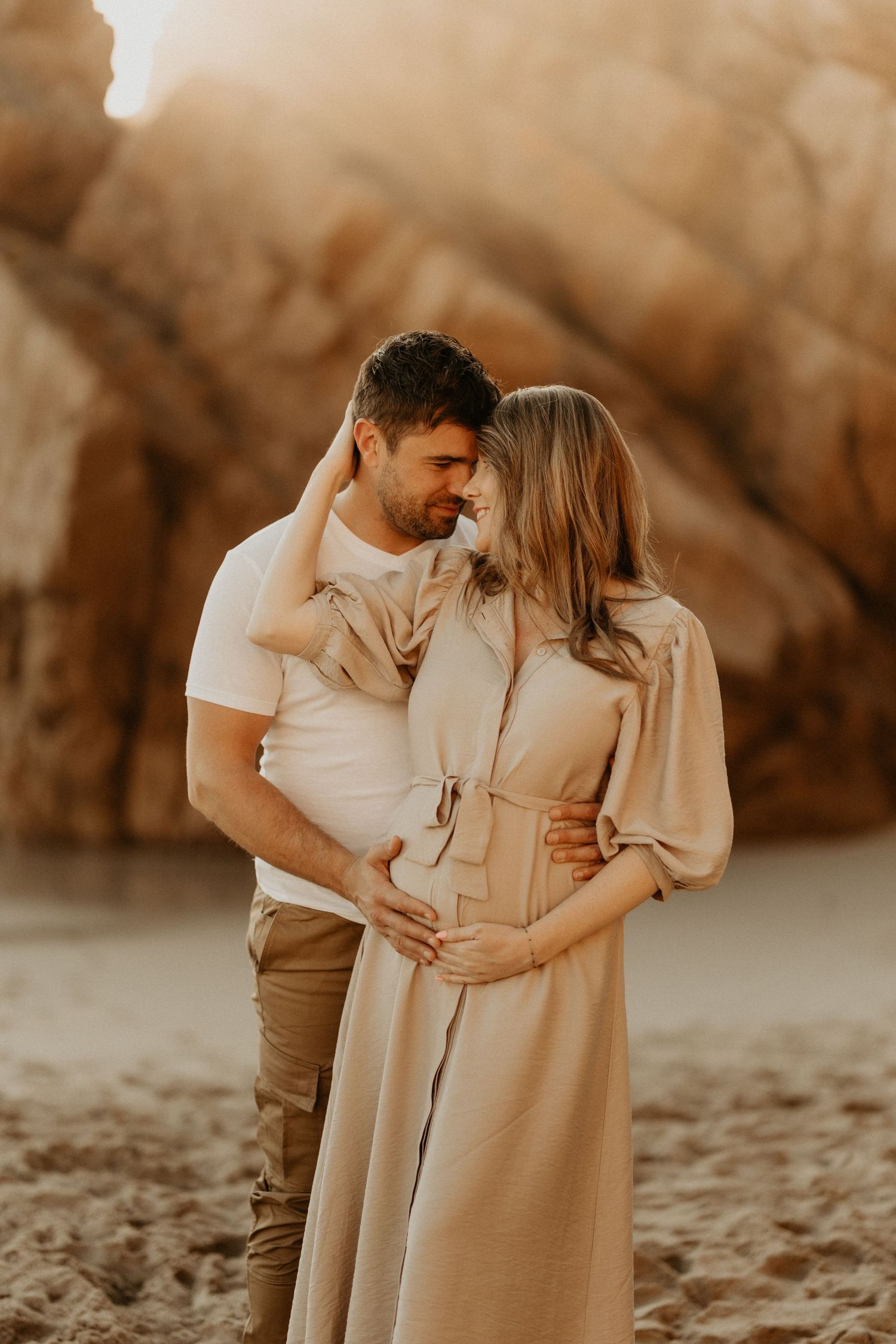 A tender moment captured between a couple on a sandy beach with rocky cliffs in the background.