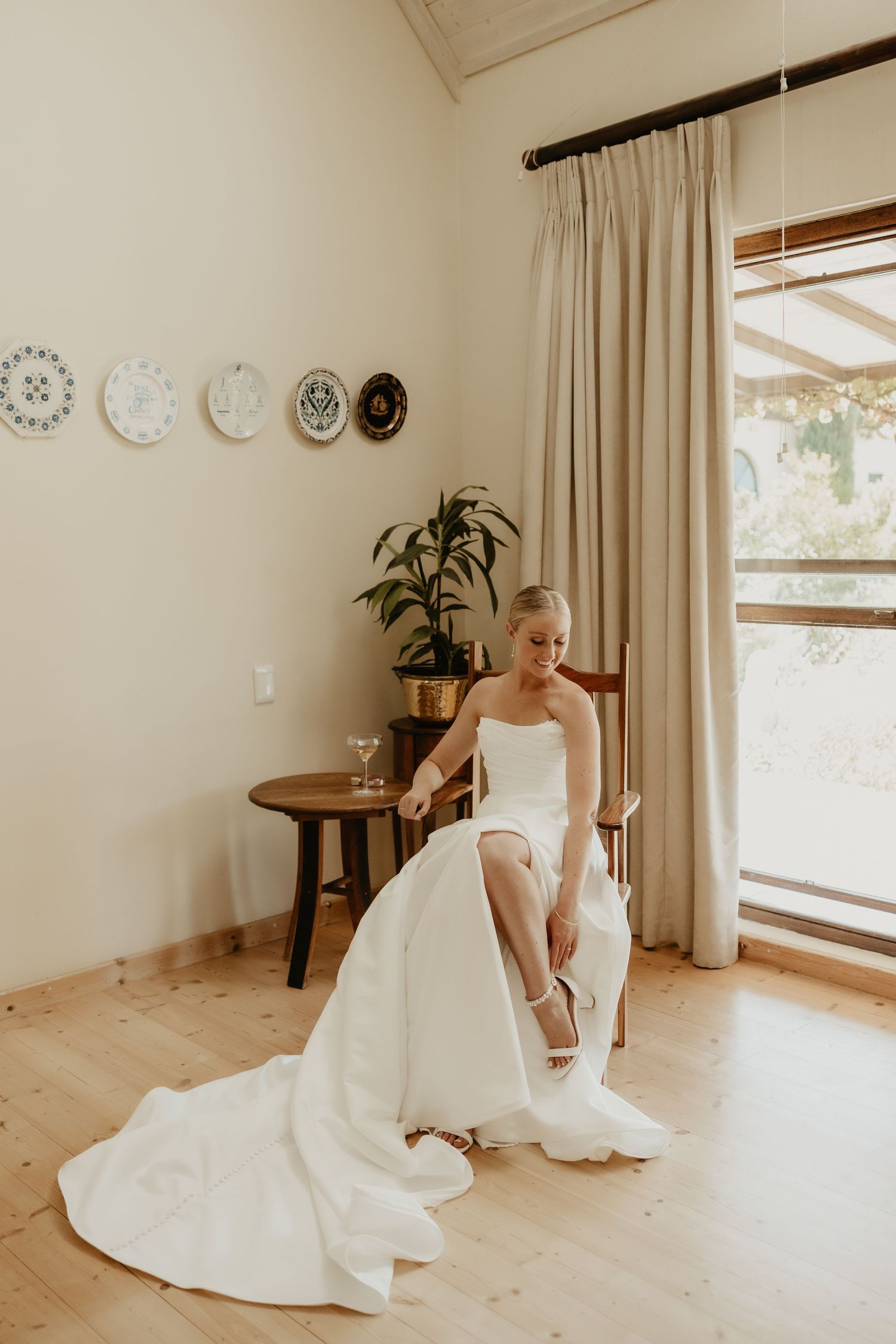 Bride in strapless white wedding gown sitting on wooden chair adjusting her heels in a bright, warmly decorated room.