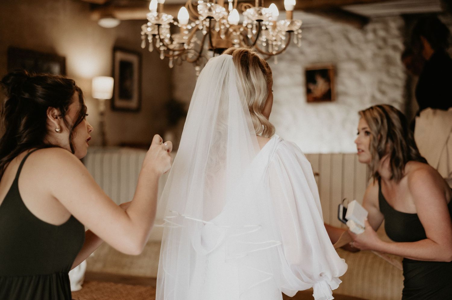 Wedding preparation moment with bridesmaids in black dresses helping bride under crystal chandelier.