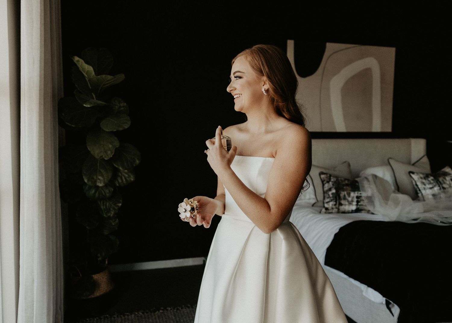 A bride in a white strapless wedding dress laughs joyfully while getting ready in a bedroom.