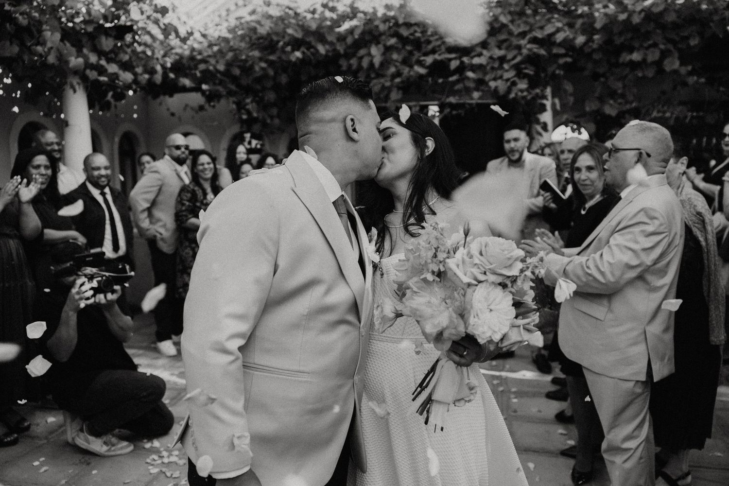 A celebratory wedding kiss surrounded by guests in an outdoor garden setting.