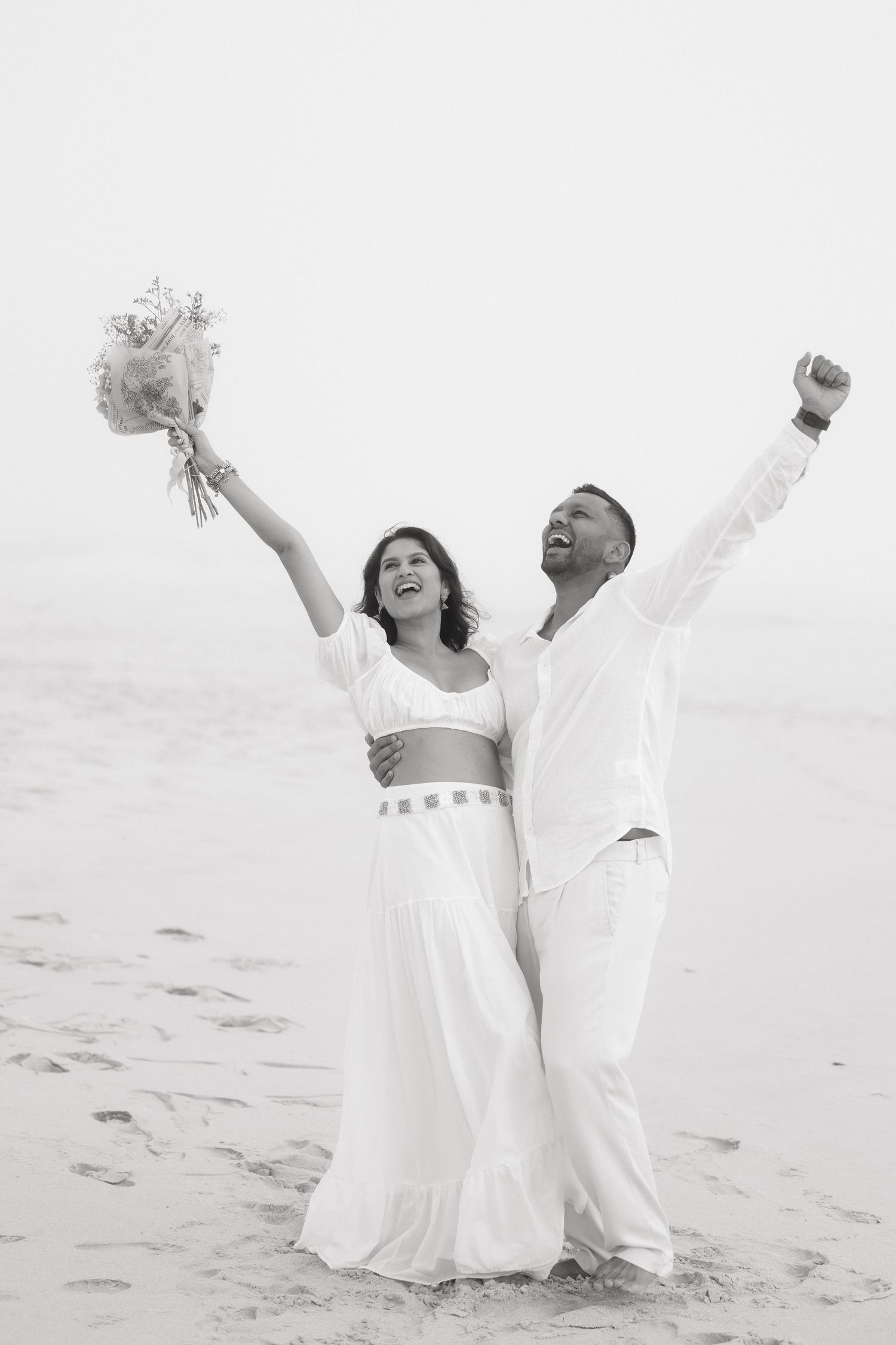 Black and white photo of joyful couple in white celebrating on beach, woman holding bouquet, both raising arms in triumph.