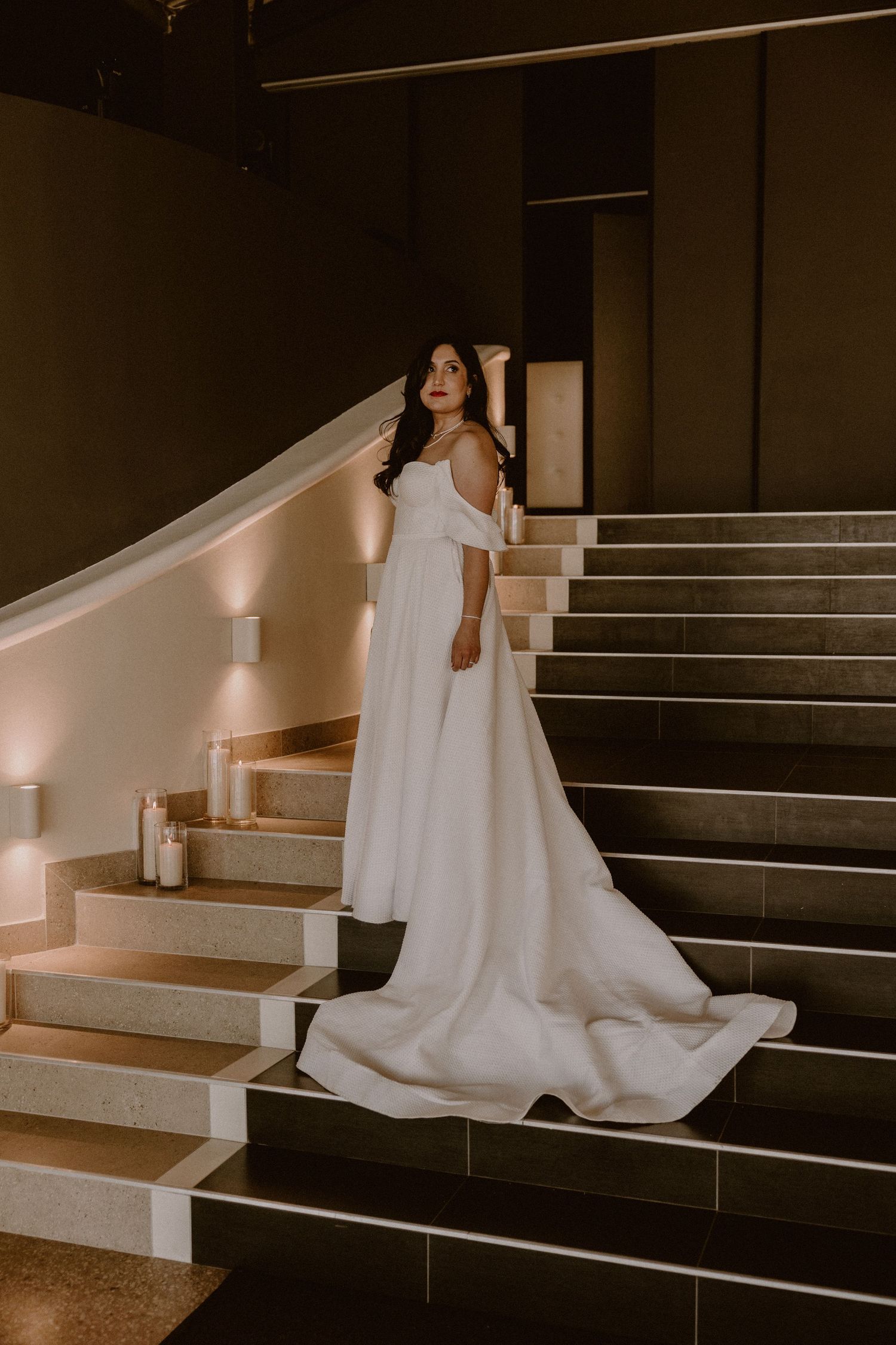 Bride in flowing white dress poses on elegant indoor staircase with dramatic lighting.