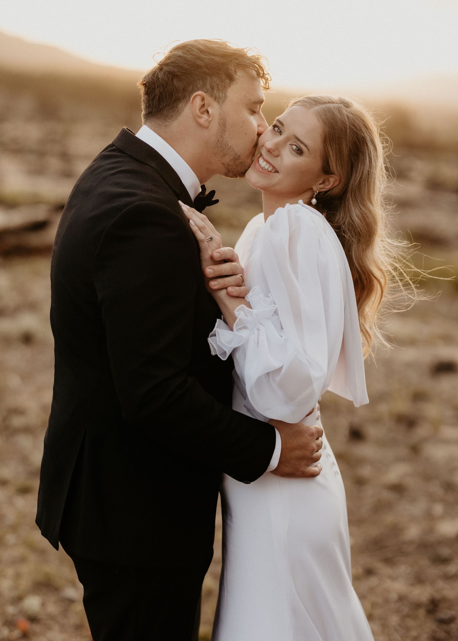 Romantic wedding photo of couple embracing in golden sunlight with bride in flowing white dress and groom in tuxedo.