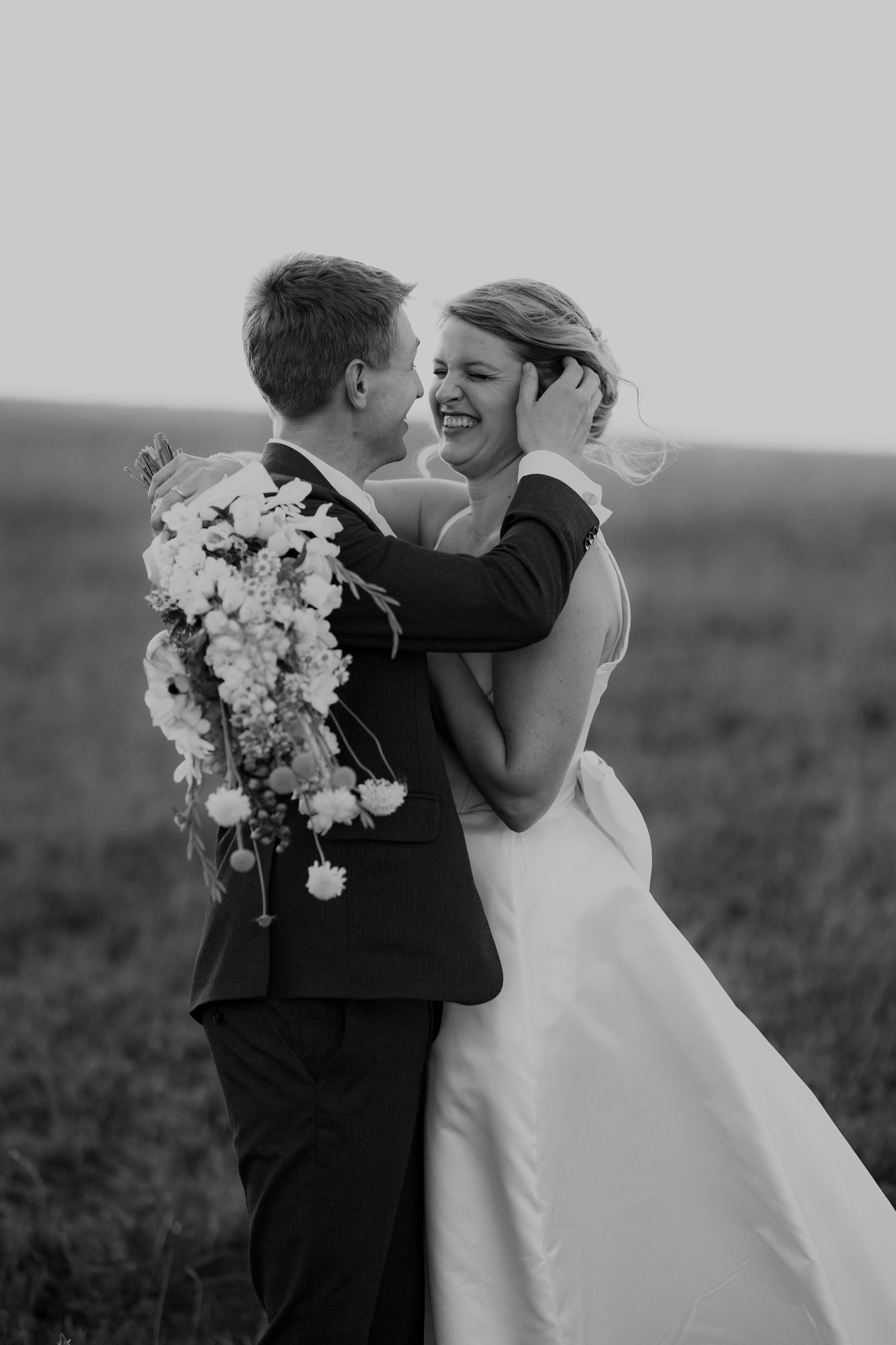 A joyful wedding couple shares an intimate black and white moment together while holding a white floral bouquet.