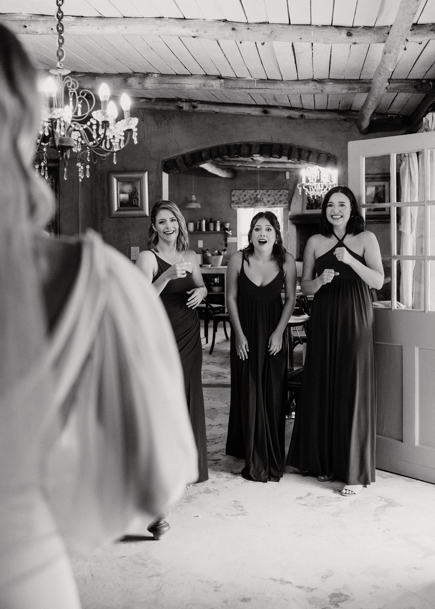 Bridesmaids in black dresses reacting with joy in a rustic room with chandelier.