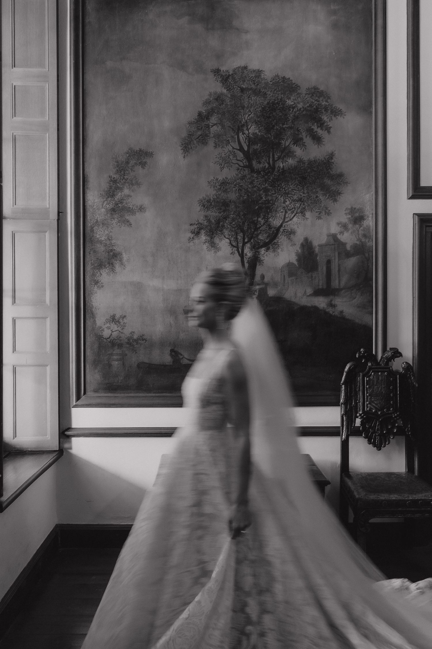 Black and white photo of a bride in a flowing wedding gown and veil, moving gracefully before a large landscape mural.