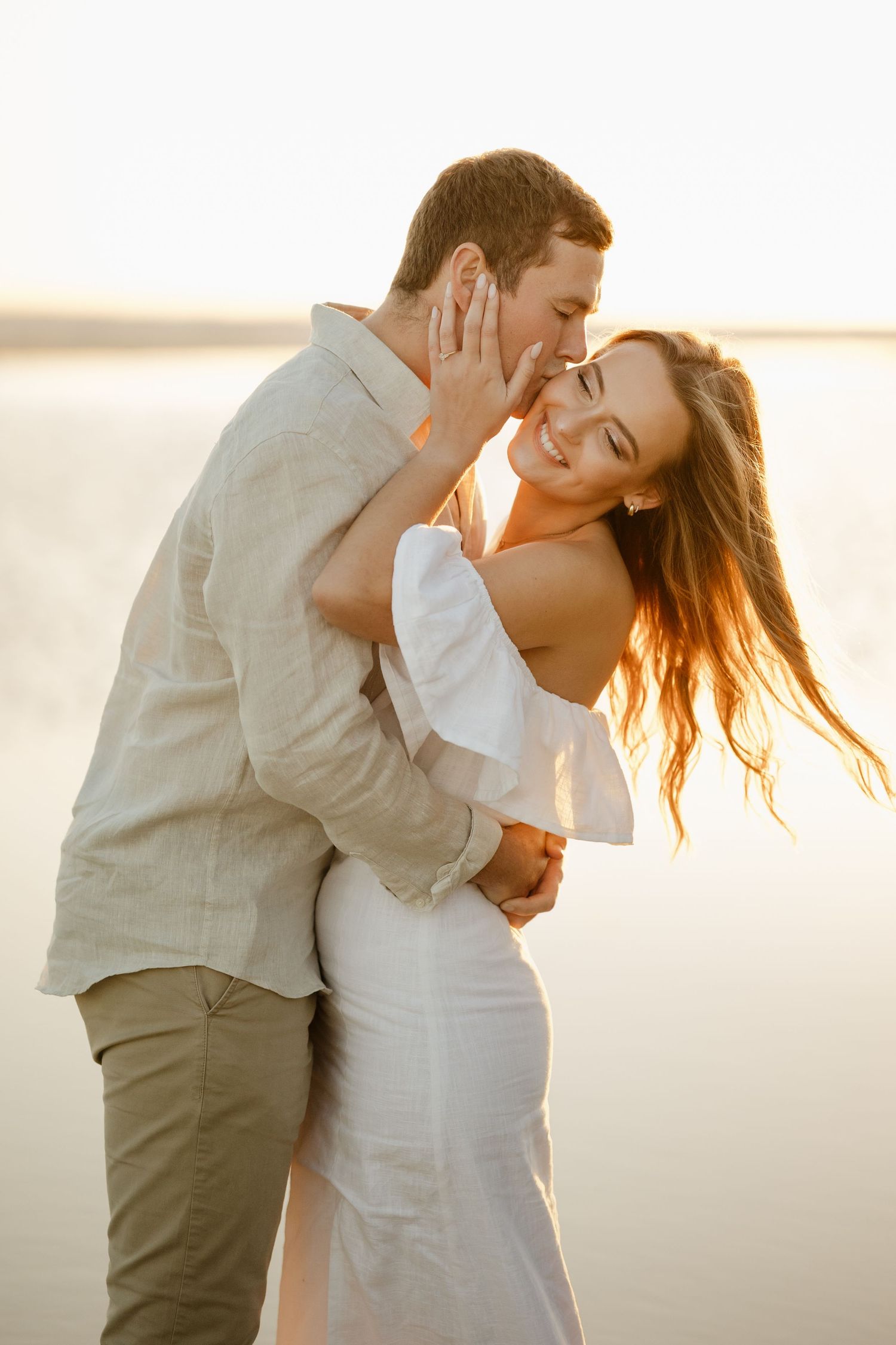 Couple embracing at sunset by the water, woman in white dress smiling as man kisses her cheek.