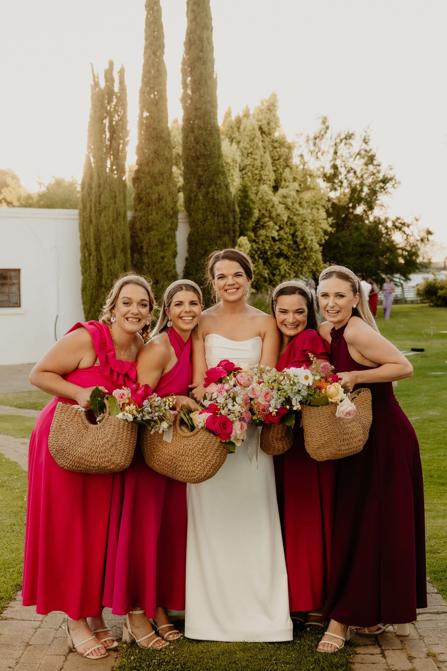 Bridal party in red bridesmaid dresses and white wedding gown pose together with woven baskets and flowers outdoors.