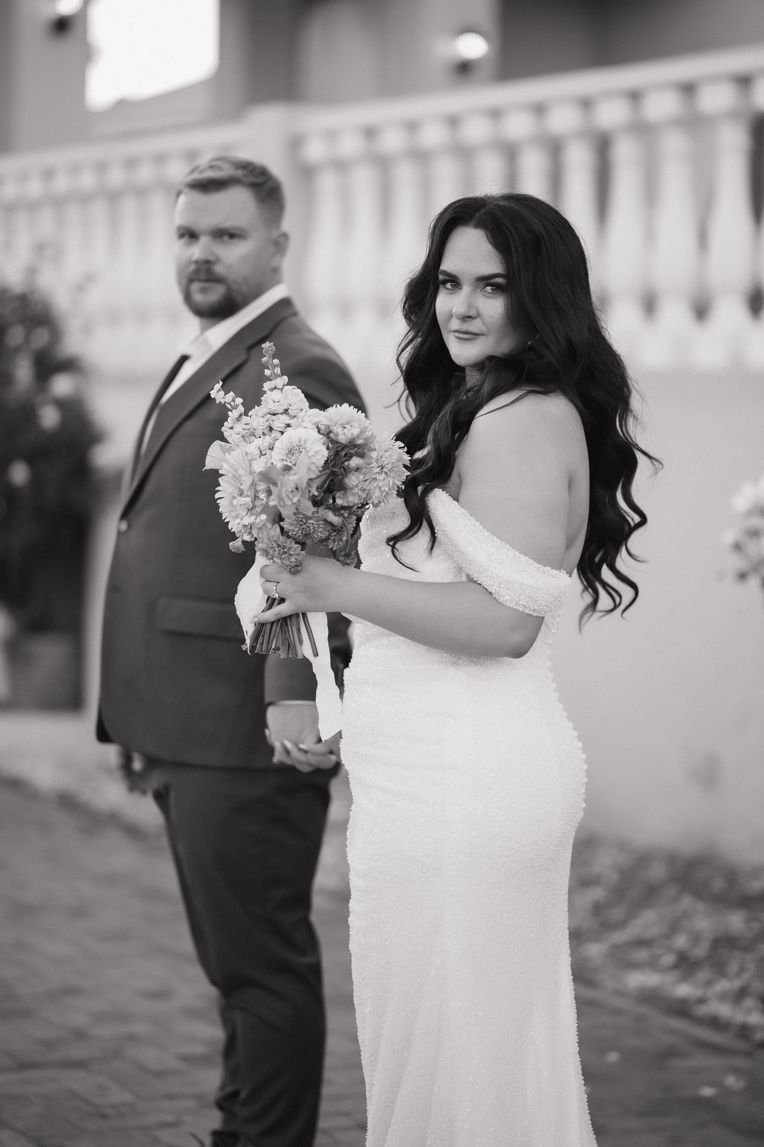 Black and white wedding photo of bride in white gown holding bouquet, standing with groom in suit outdoors.