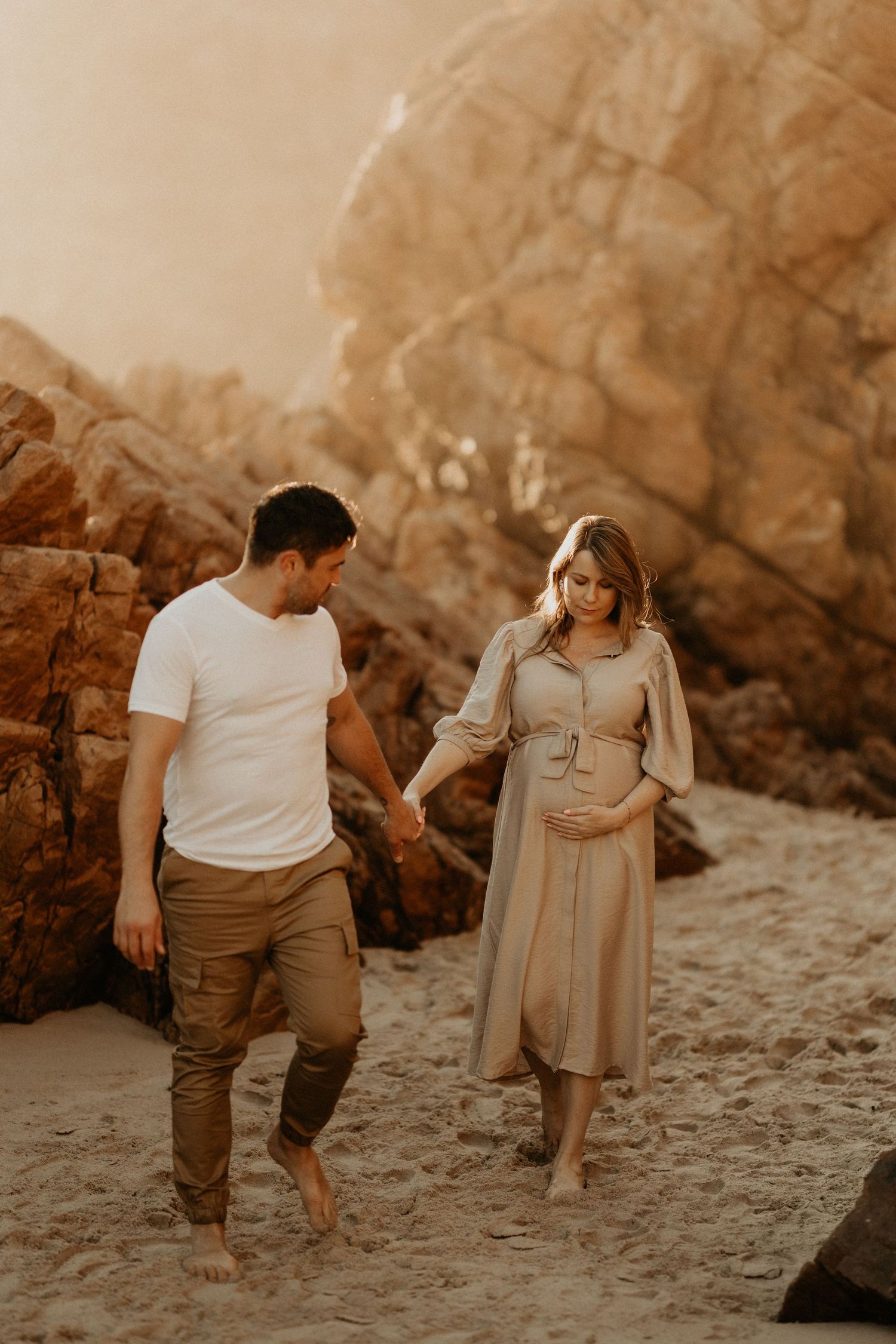 An expectant couple walks barefoot on beach sand with golden cliffs in the background.