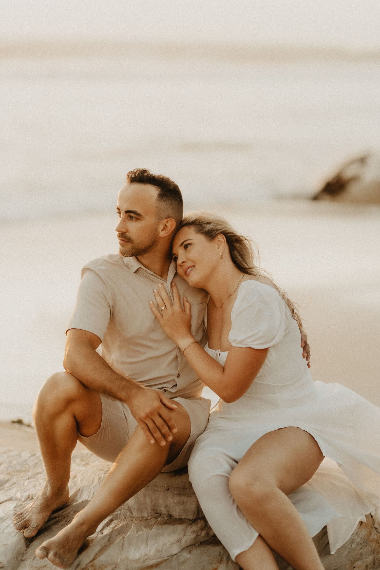 A couple in light colored outfits sits intimately together on coastal rocks during golden hour.