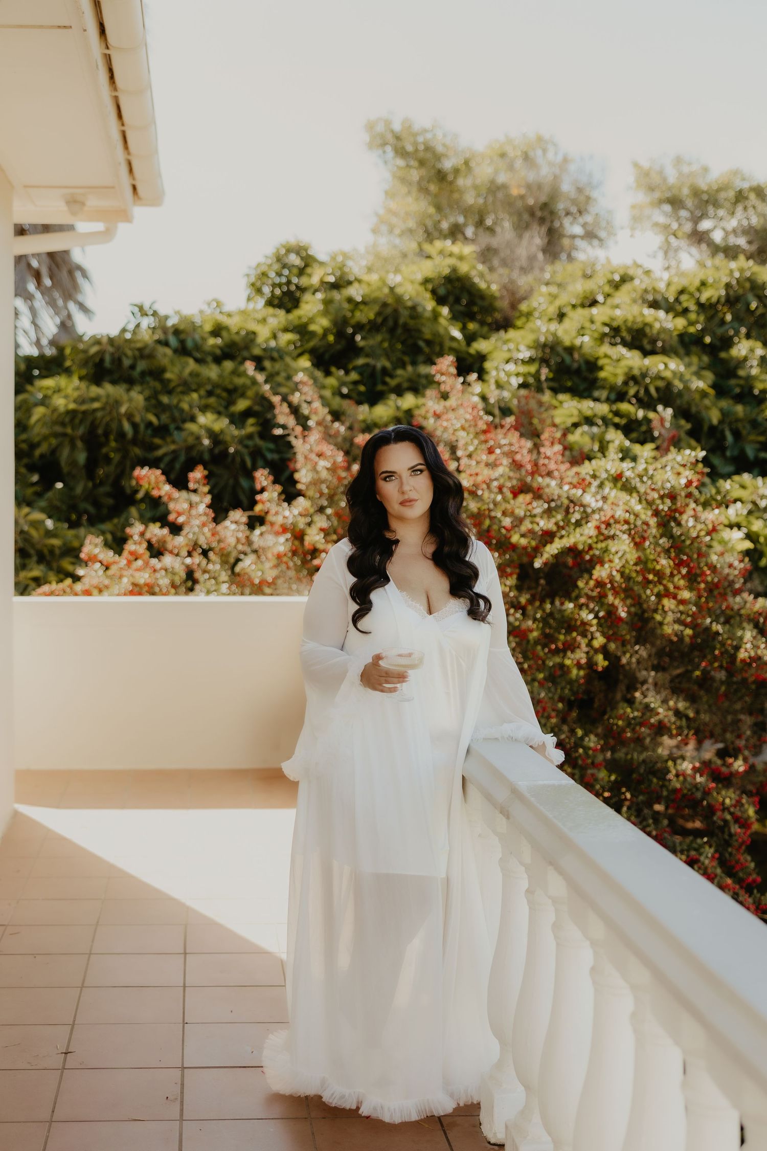 Bride in white bridal robe holding a glass on a sunlit balcony surrounded by lush green and flowering trees.