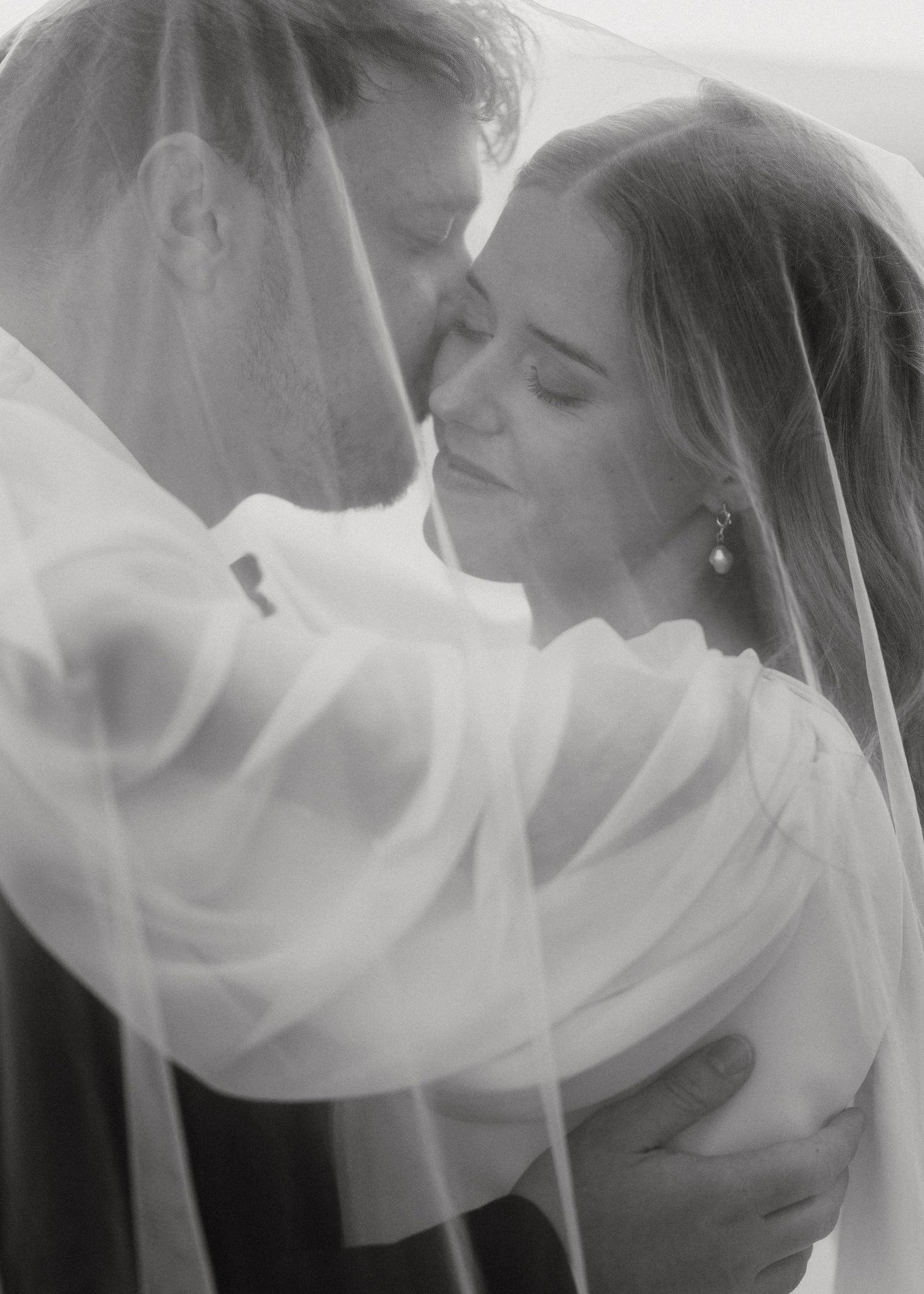An intimate wedding portrait shows a couple sharing a tender moment beneath a billowing veil.
