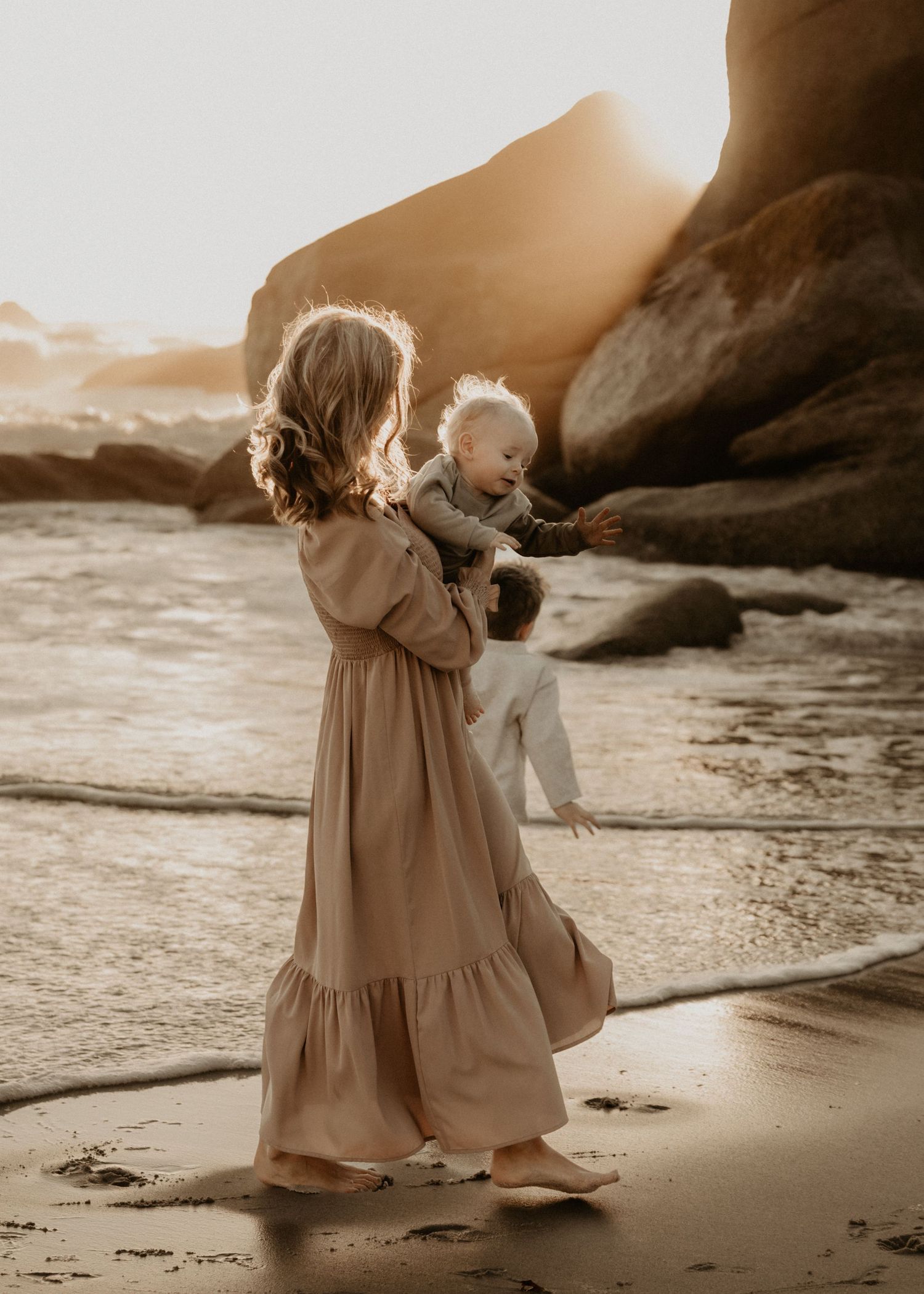 Person in flowy dress walks along beach shore at sunset with dramatic rock formations.
