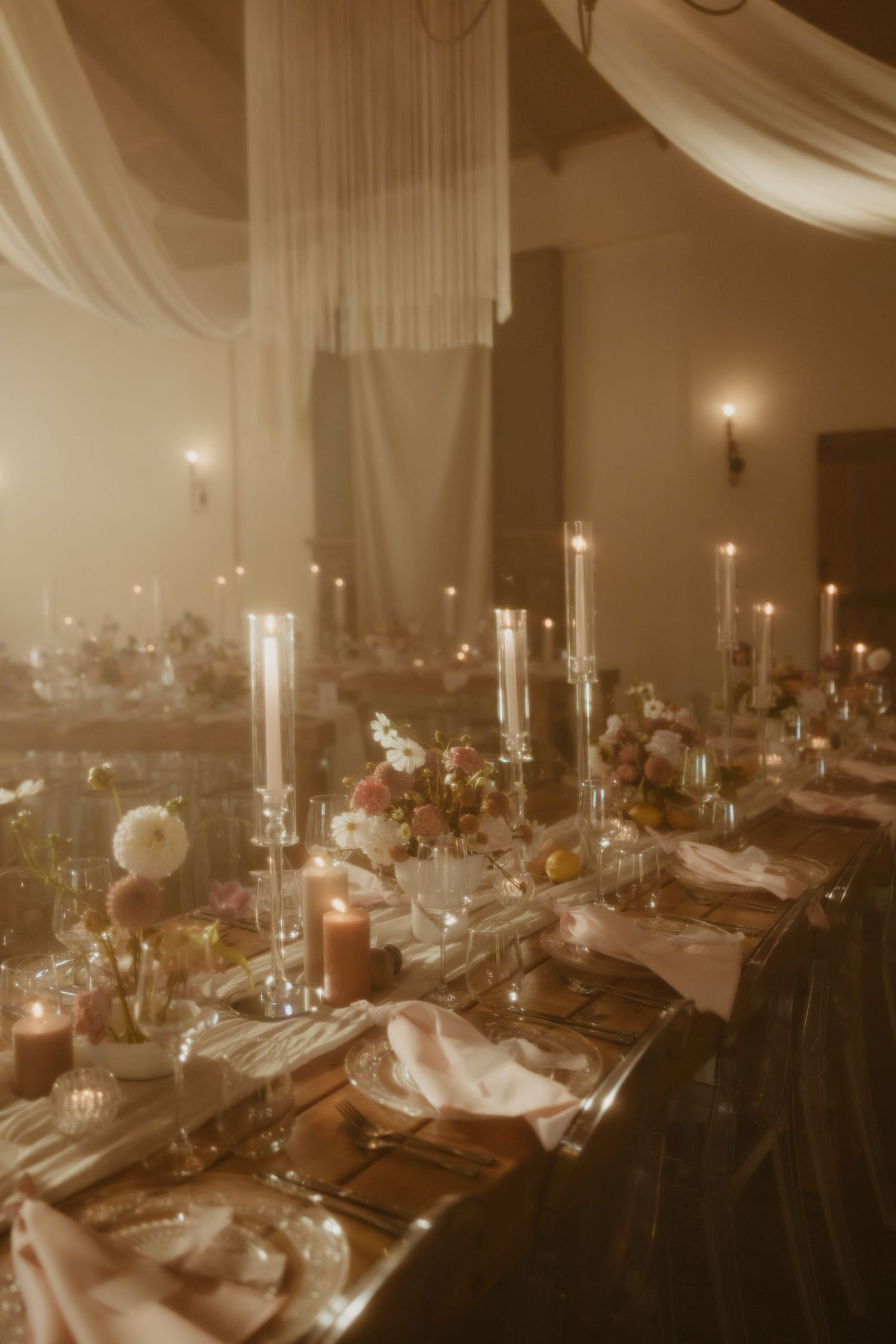 Elegant wedding reception with candlelit long tables, floral centerpieces, draped fabric, and warm golden lighting.