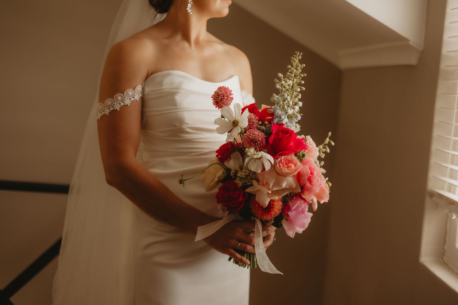 A bridal bouquet with pink and red flowers is held against a white wedding dress in natural window light.