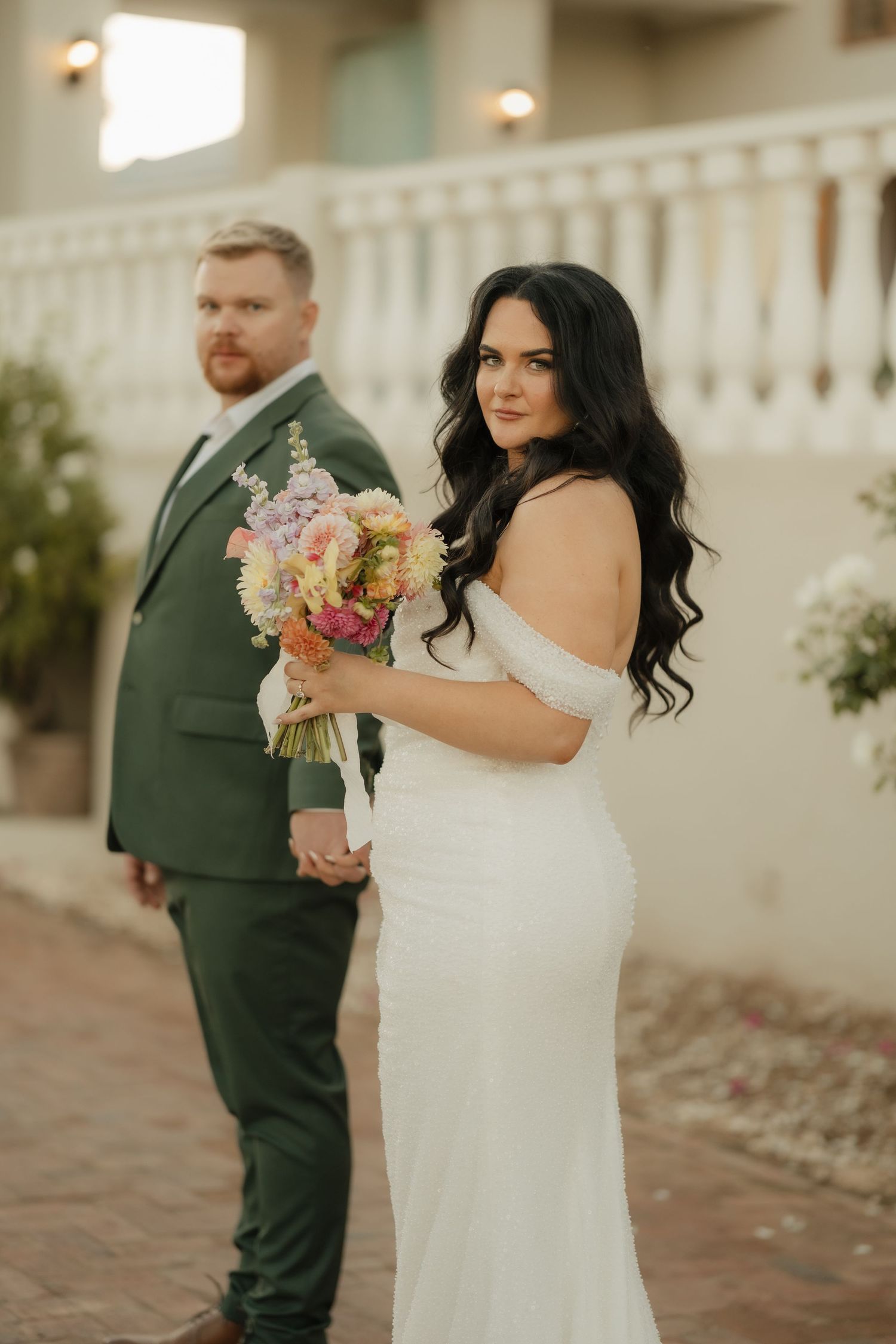 Bride in white off-shoulder gown holds colorful bouquet, groom in green suit, elegant outdoor wedding portrait.