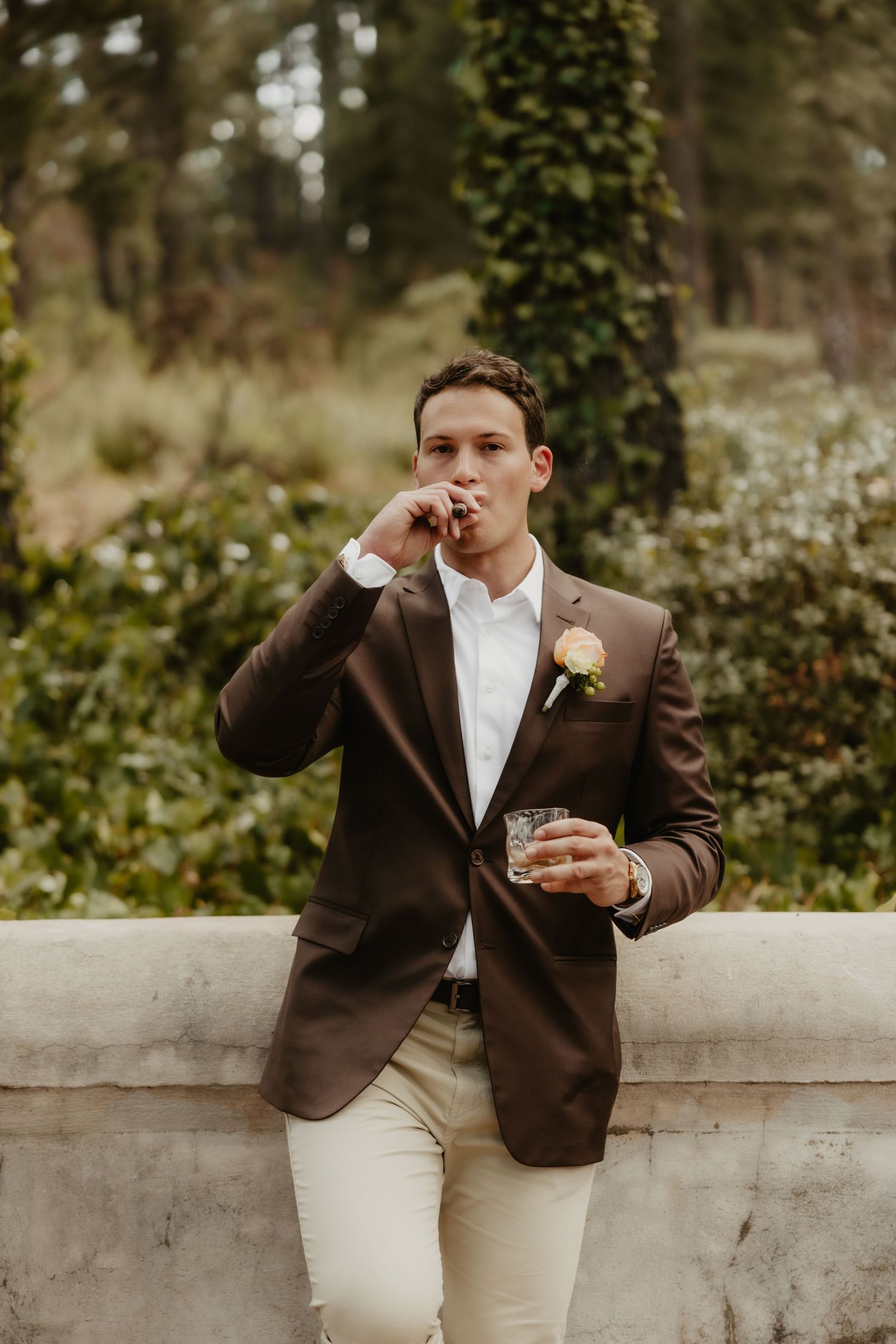 Groom in brown blazer with boutonniere smoking cigar and holding whiskey glass outdoors at wedding reception.