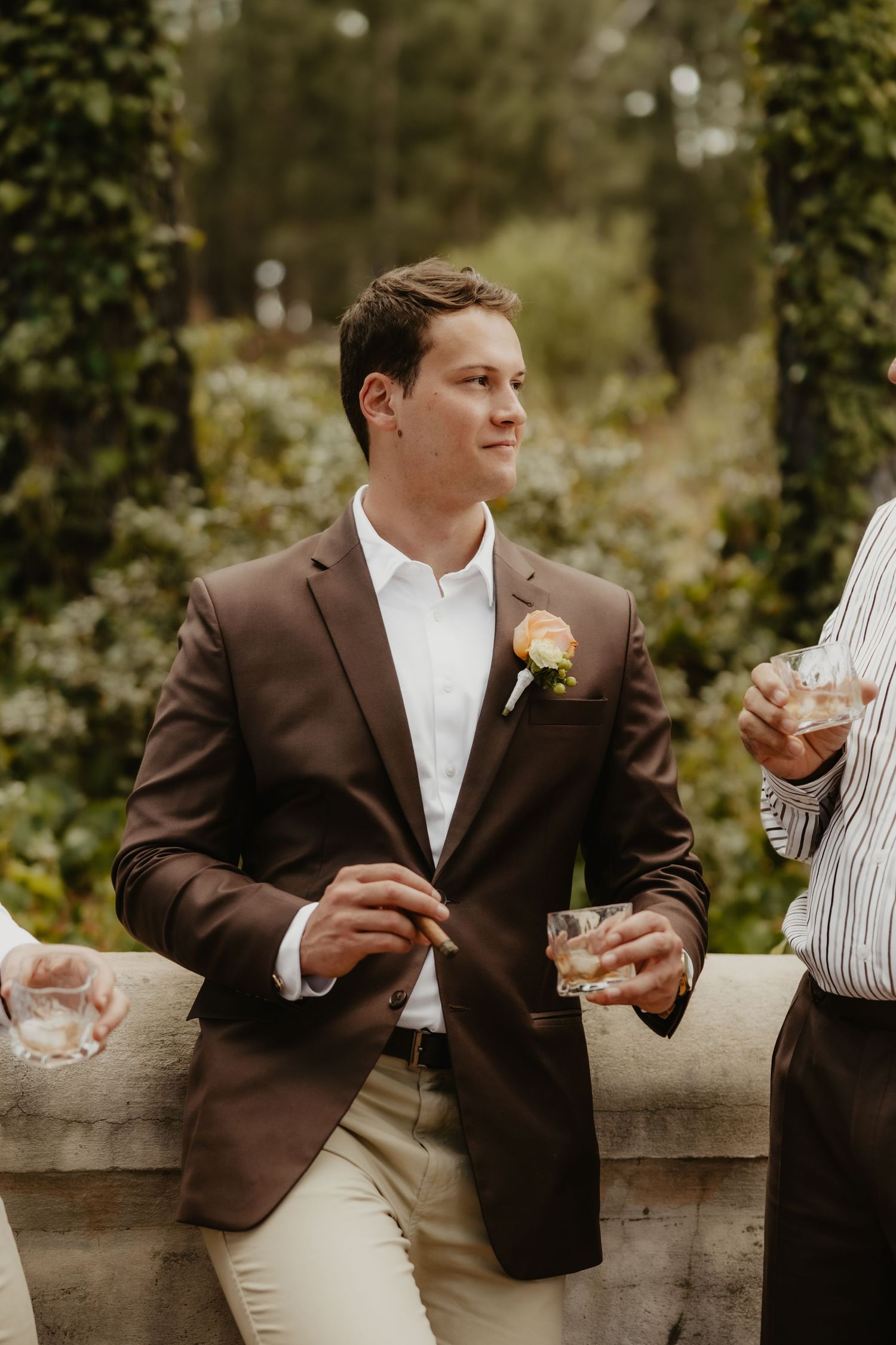 Man in brown blazer with boutonniere holds a drink at an outdoor wedding reception.