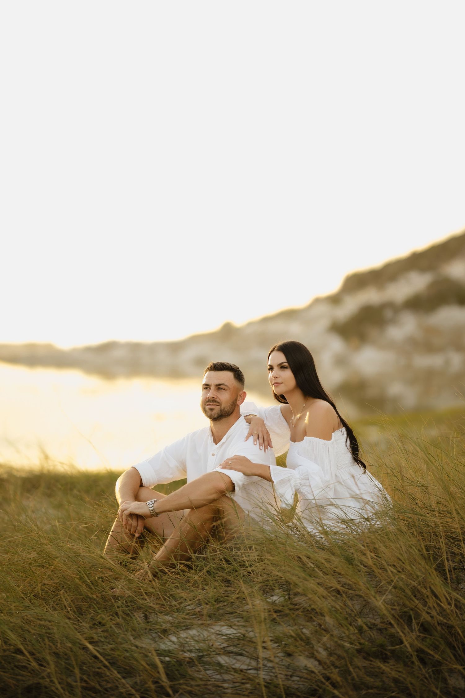 Couple in white outfits sitting together in golden grass dunes at sunset, woman leaning on mans shoulder.