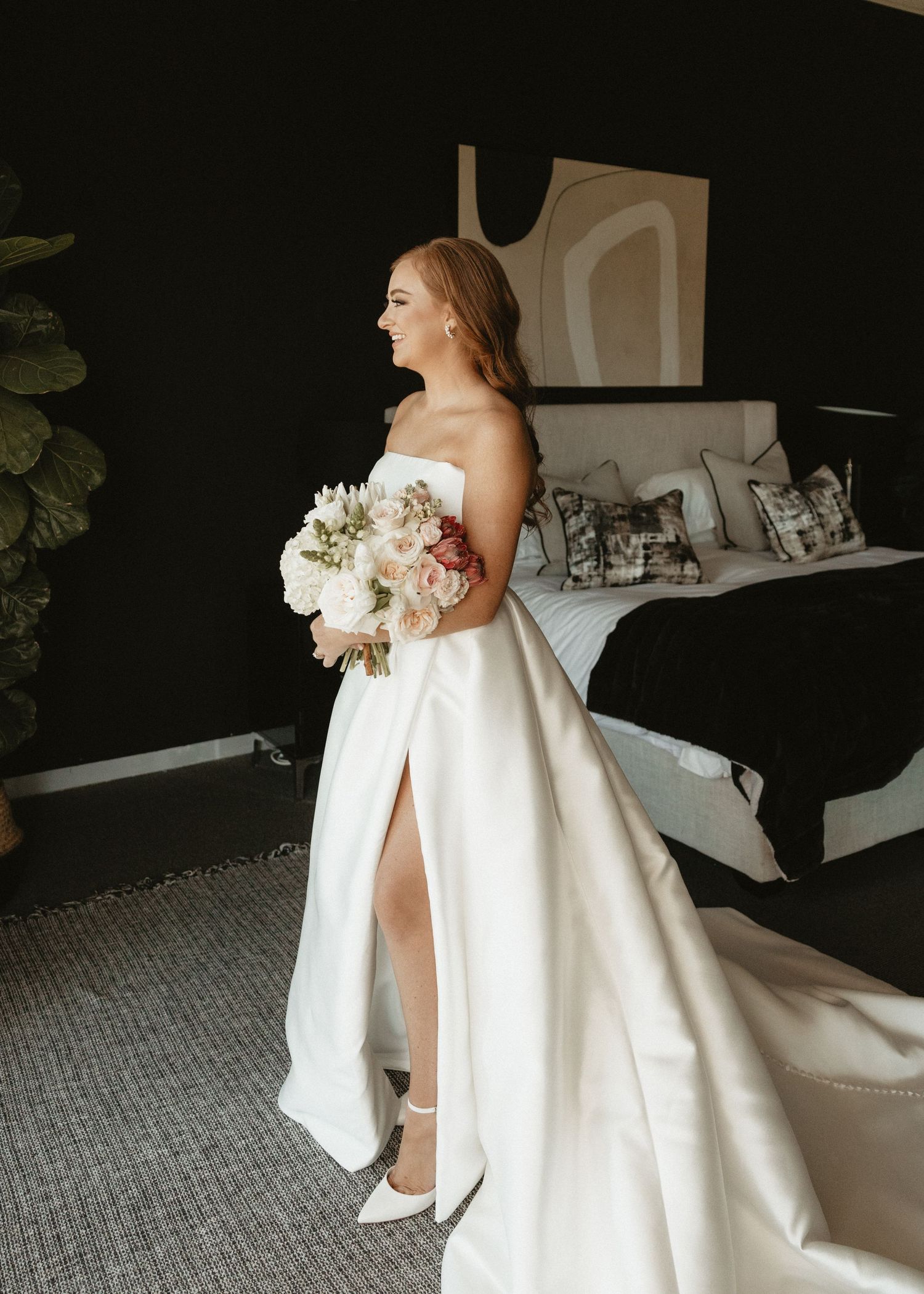 A bride holds a white floral bouquet while wearing a strapless wedding gown with a dramatic side slit.