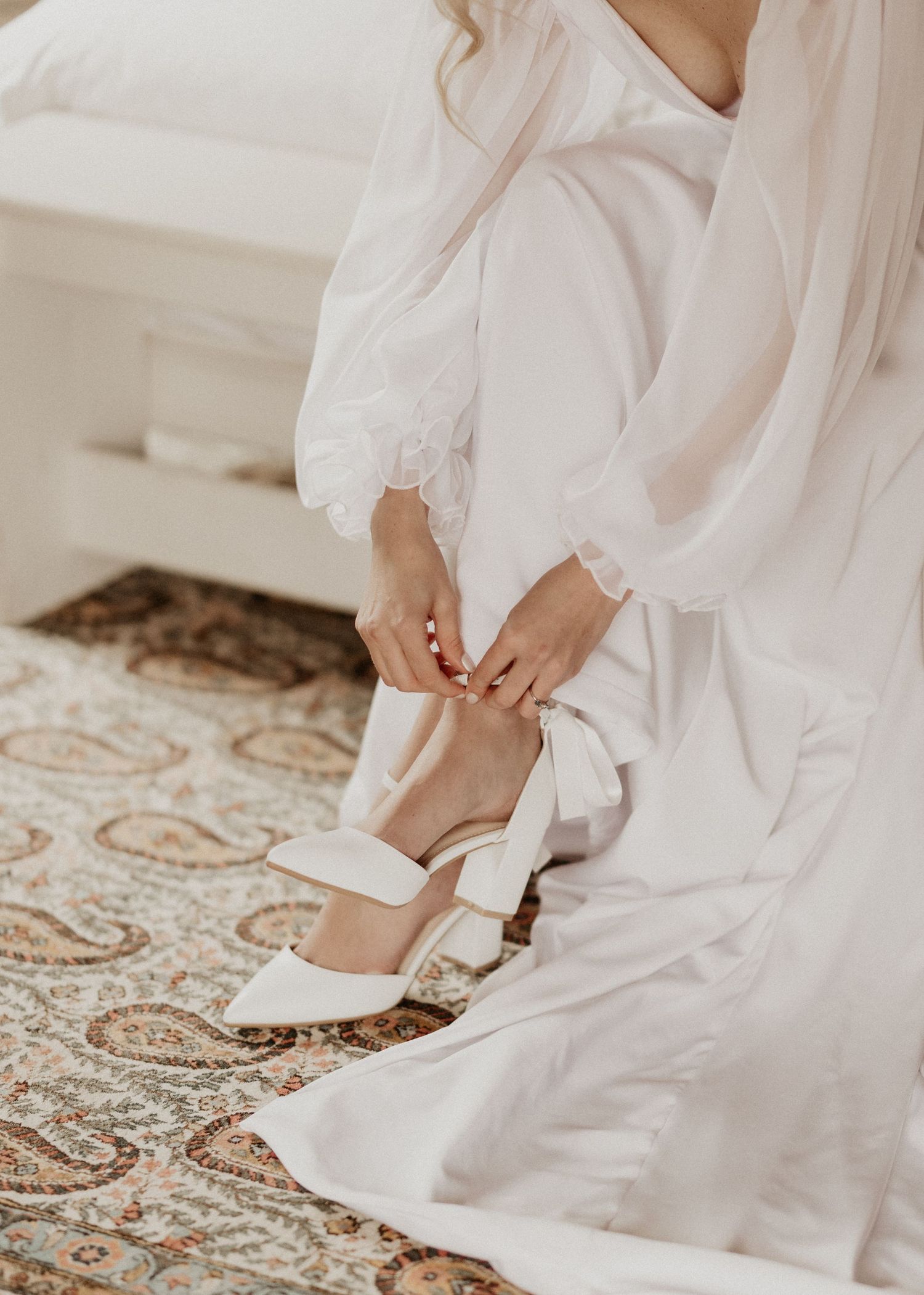 White bridal shoes being fastened on an ornate carpet next to flowing wedding dress fabric.