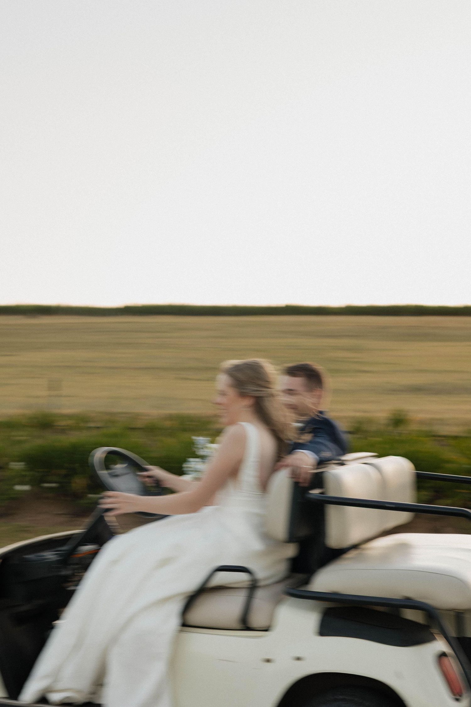 Bride in white dress and groom ride in a vintage open-top car through countryside on their wedding day.