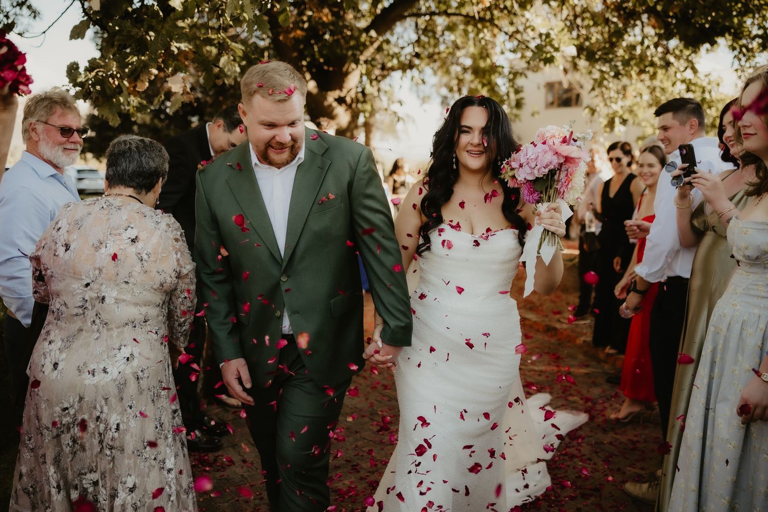 Bride and groom walk through flower petal confetti shower after outdoor wedding ceremony, guests celebrating around them.