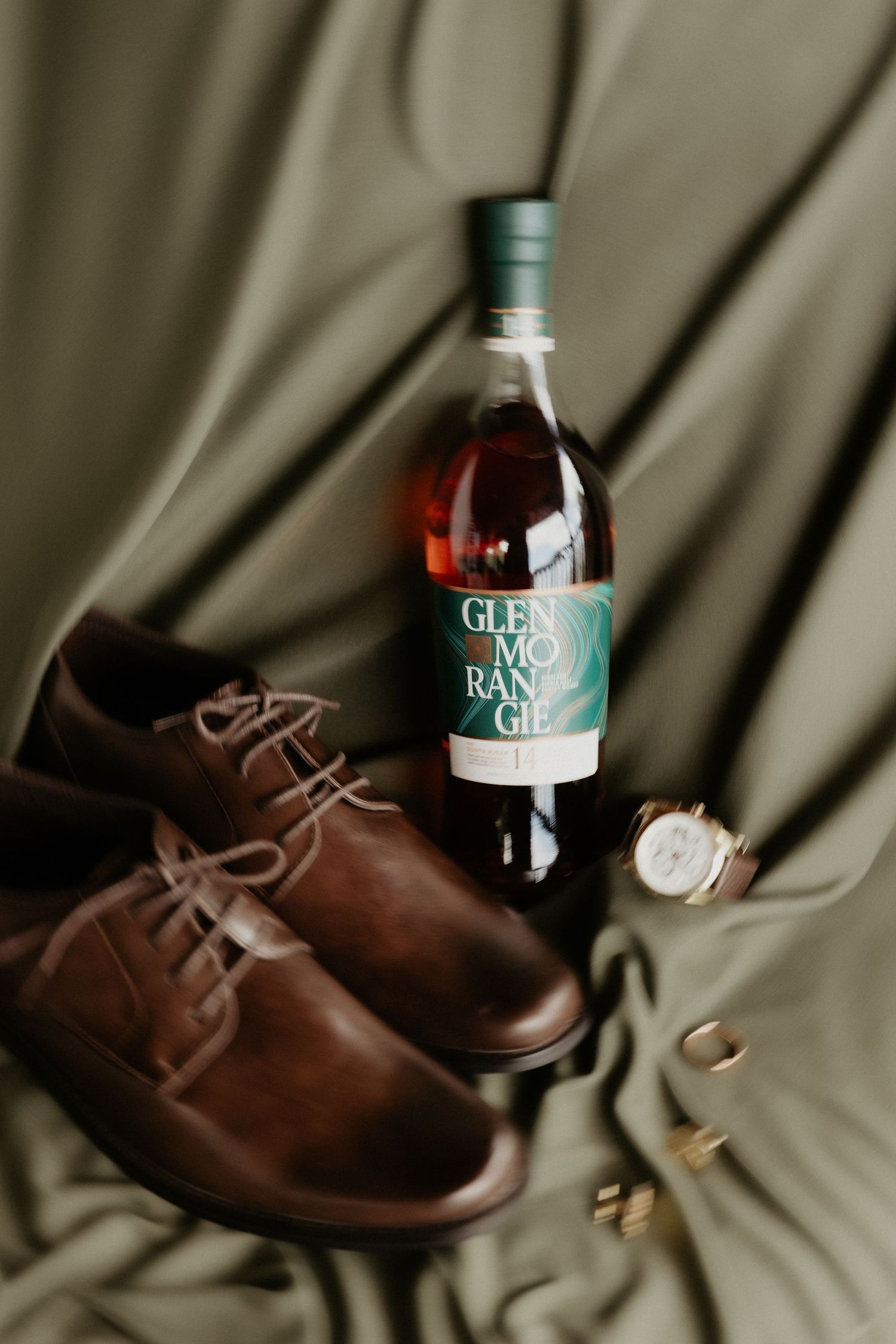 Glenmorangie whisky bottle with brown leather dress shoes, gold watch, and cufflinks on olive green fabric.