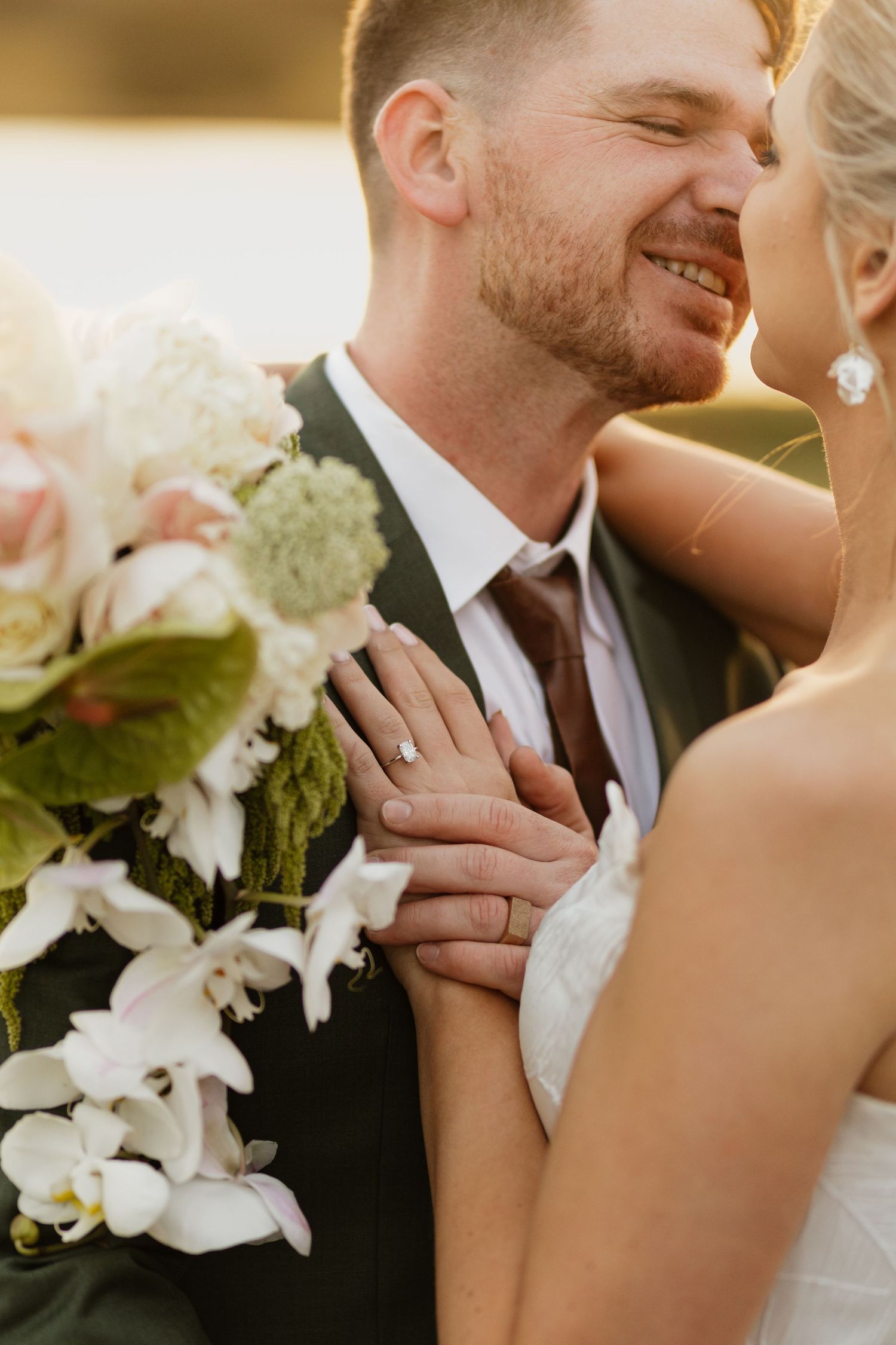 Smiling groom in dark suit holds bride close as she rests hand on his chest, showcasing engagement ring and bridal bouquet.