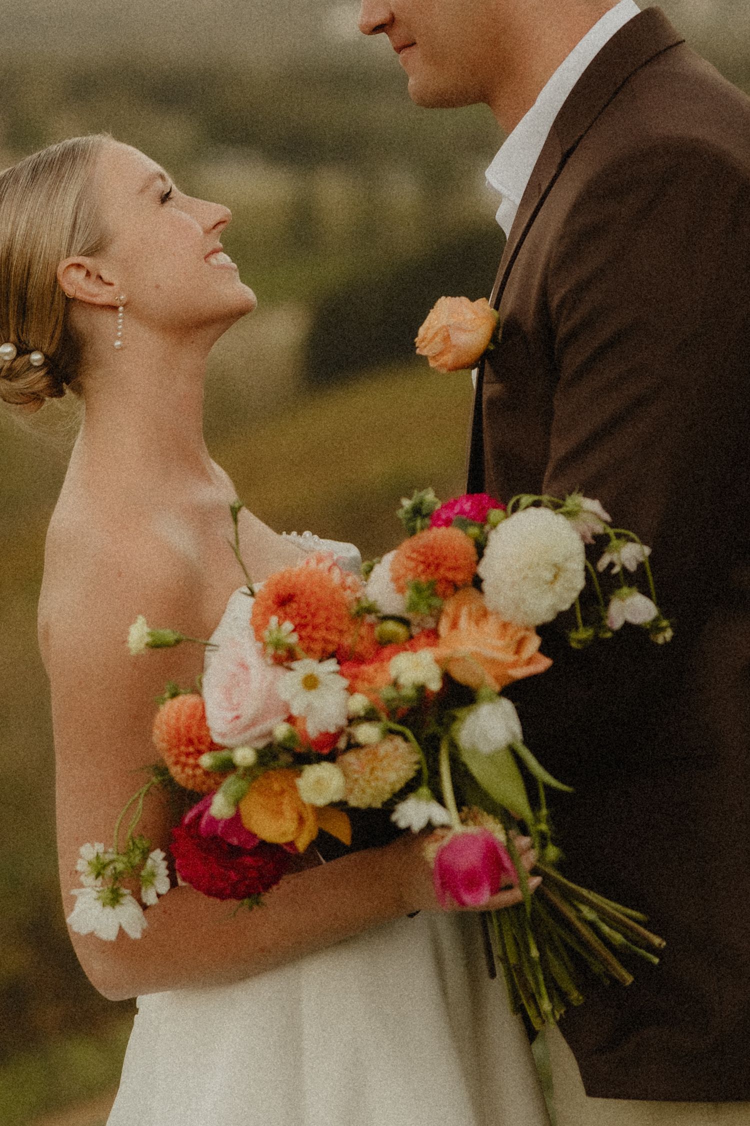 Bride laughing joyfully holding colorful bouquet of dahlias and wildflowers while gazing at groom in brown suit outdoors.