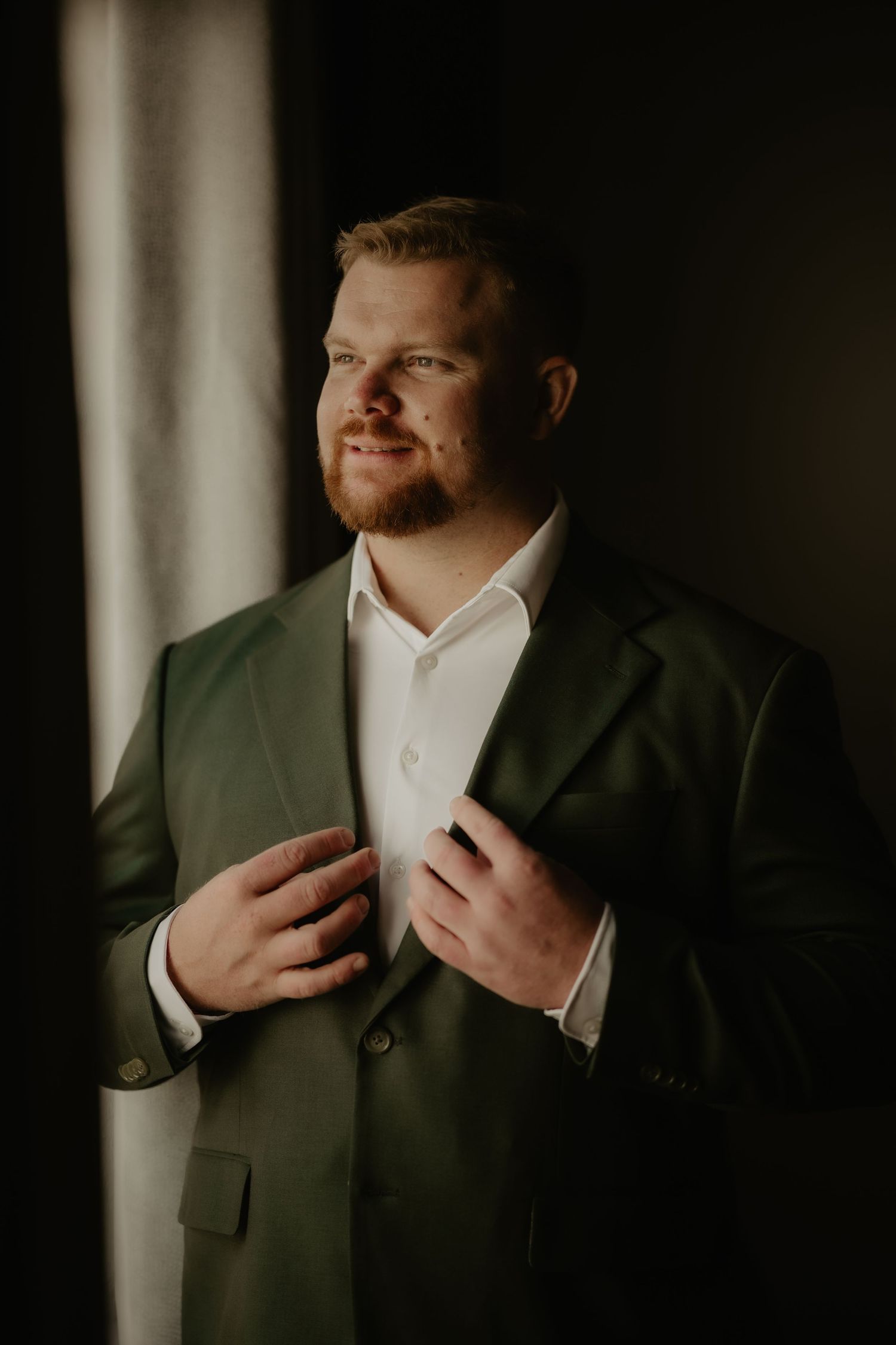 A man in a green suit jacket adjusts his lapel while standing near a window in soft, moody lighting.