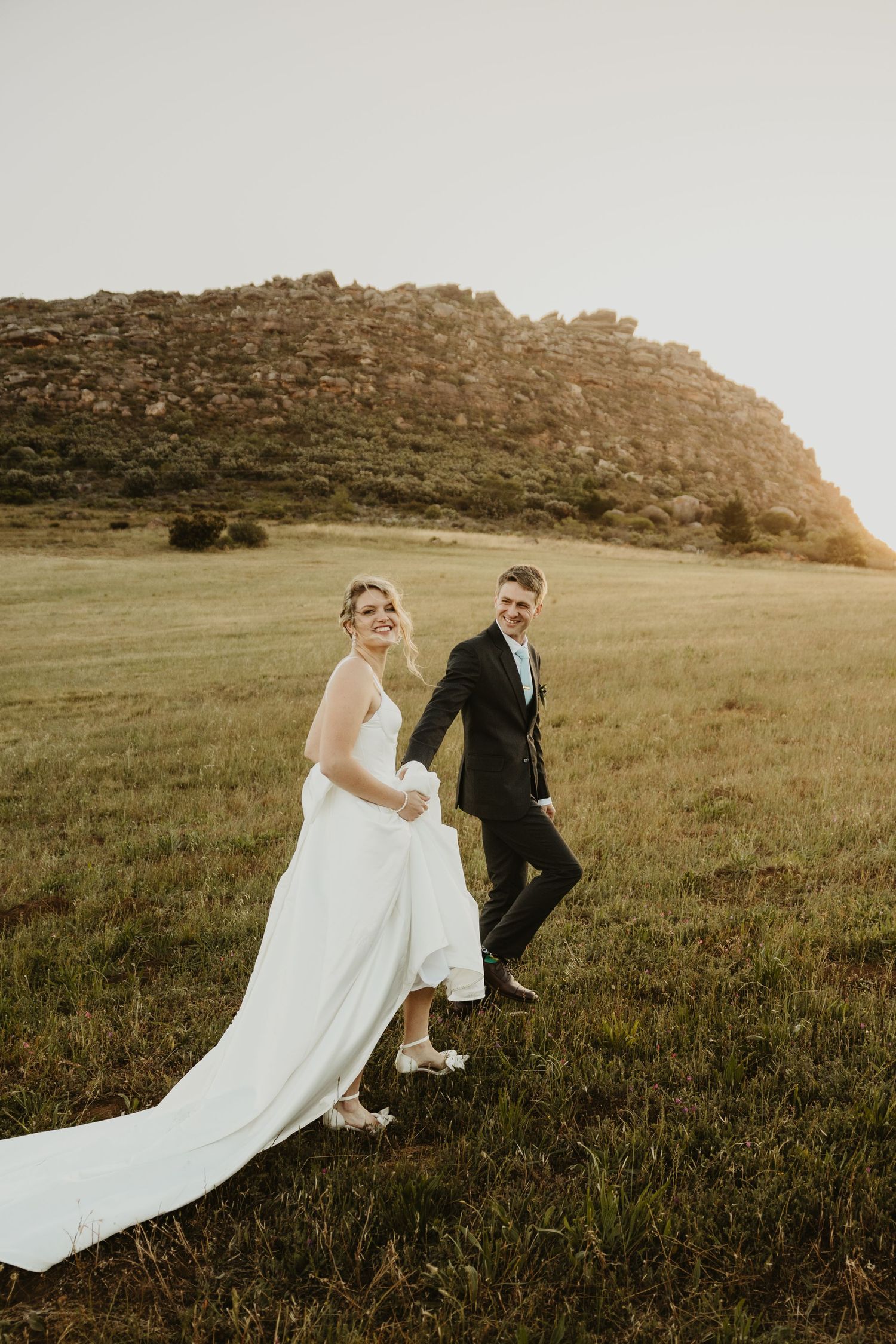 A couple in wedding attire walks through a scenic grassy meadow at sunset with mountains in the background.
