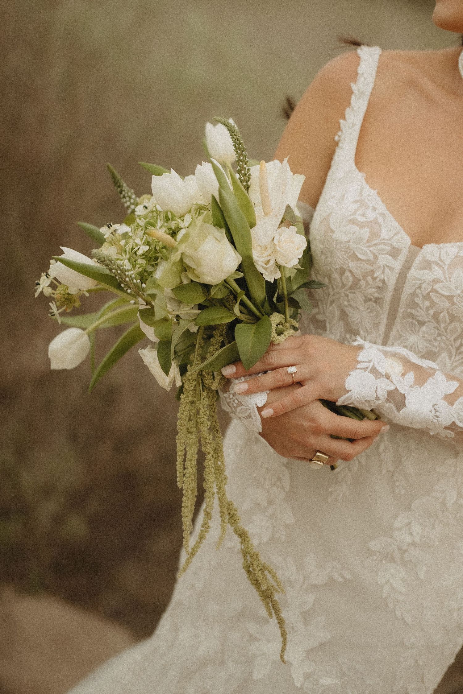Bride in lace gown holding elegant white tulip bouquet with greenery and trailing amaranthus.