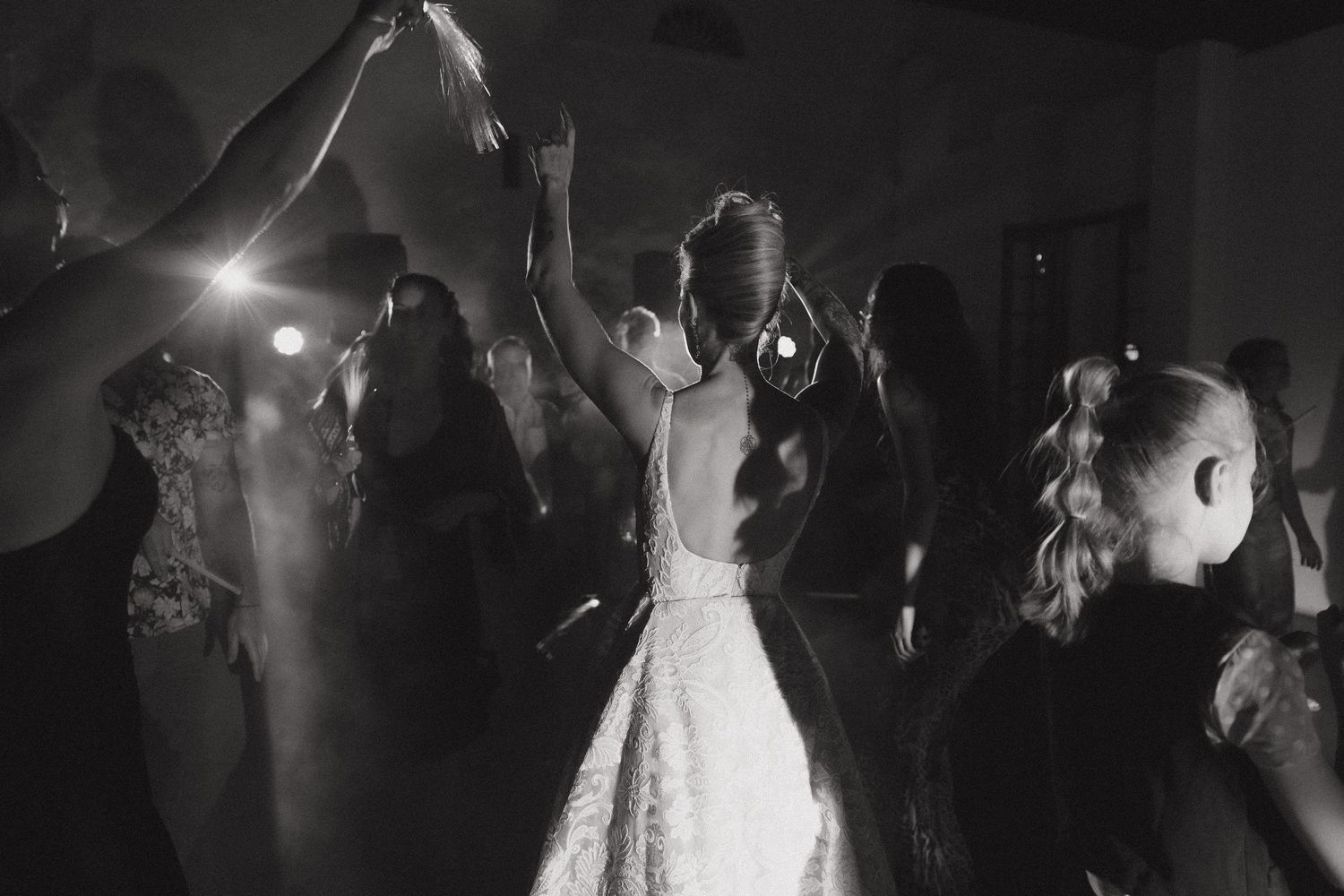 Black and white photo of a bride dancing with guests at a wedding reception, arms raised in celebration.
