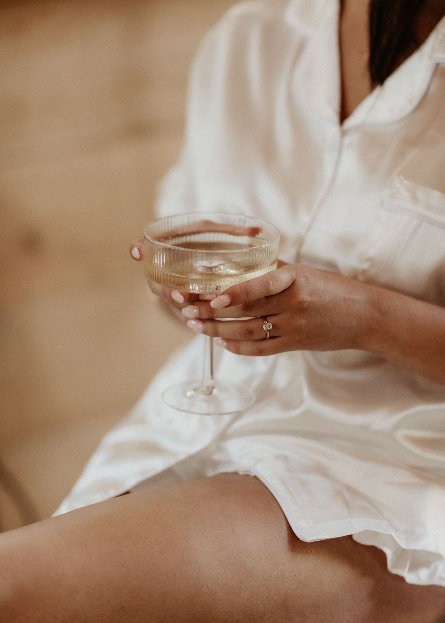 Hands holding a champagne glass while wearing a white silk robe with an elegant ring on display.