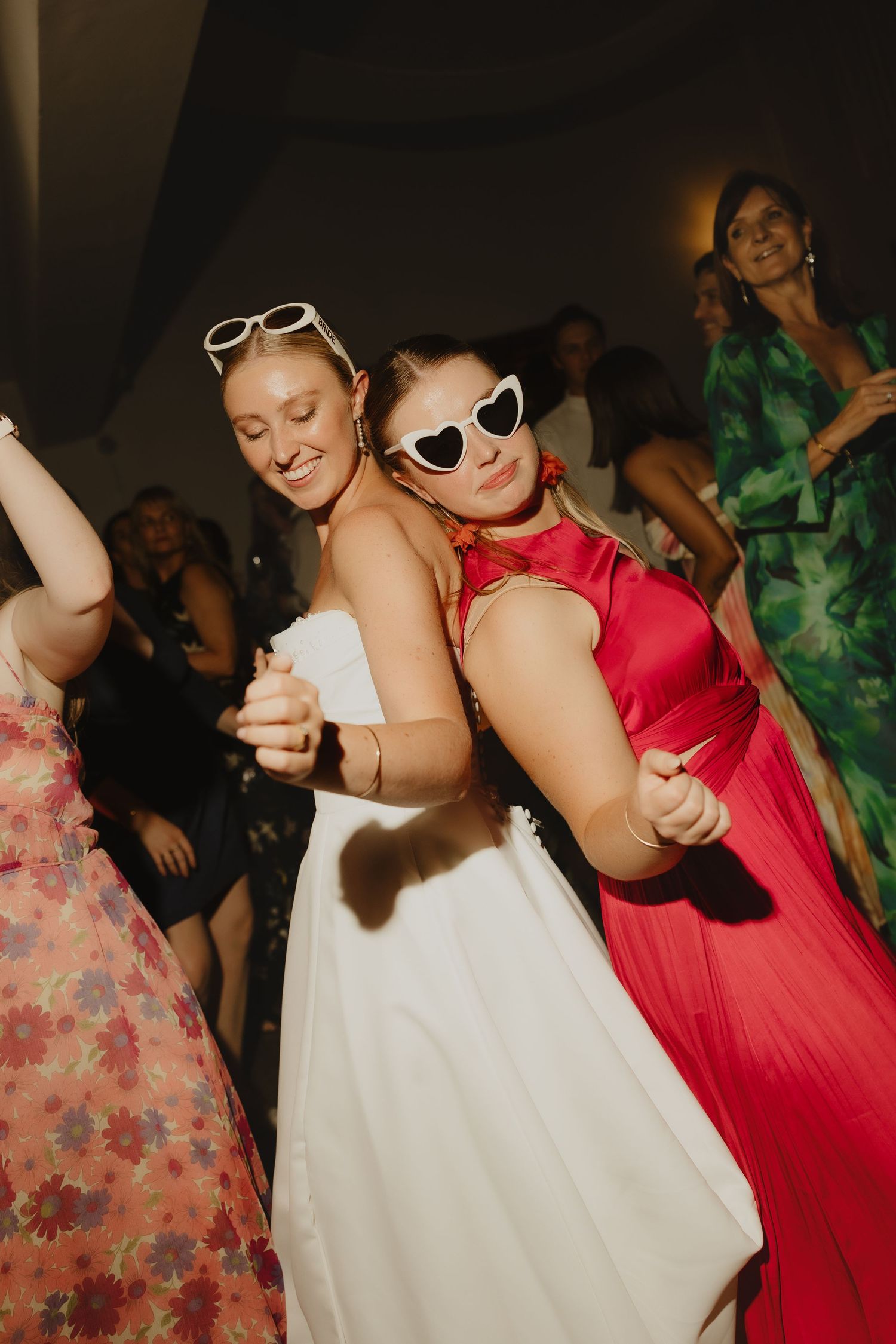 Two women dancing at a wedding reception, one in white bridal gown and one in red dress wearing heart-shaped sunglasses.
