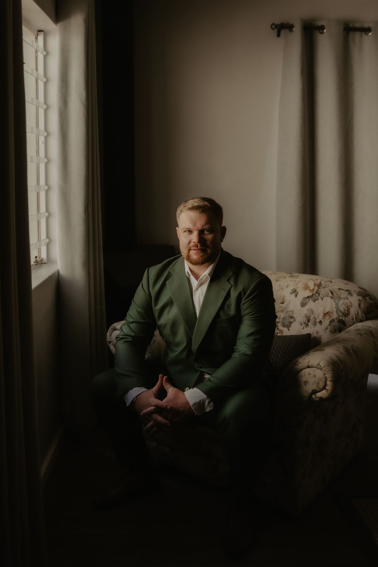 Man in green suit sitting on floral chair by window in moody indoor portrait.