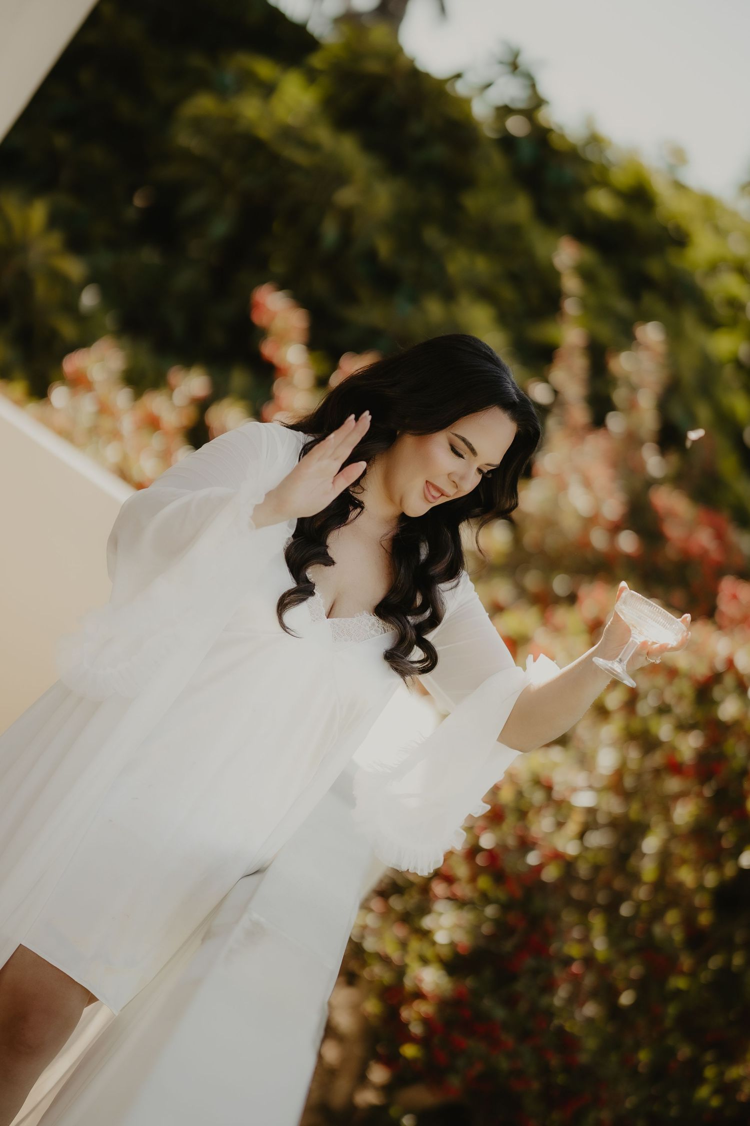 A bride in a white tulle robe reads a note outdoors, surrounded by lush greenery and blooming flowers.