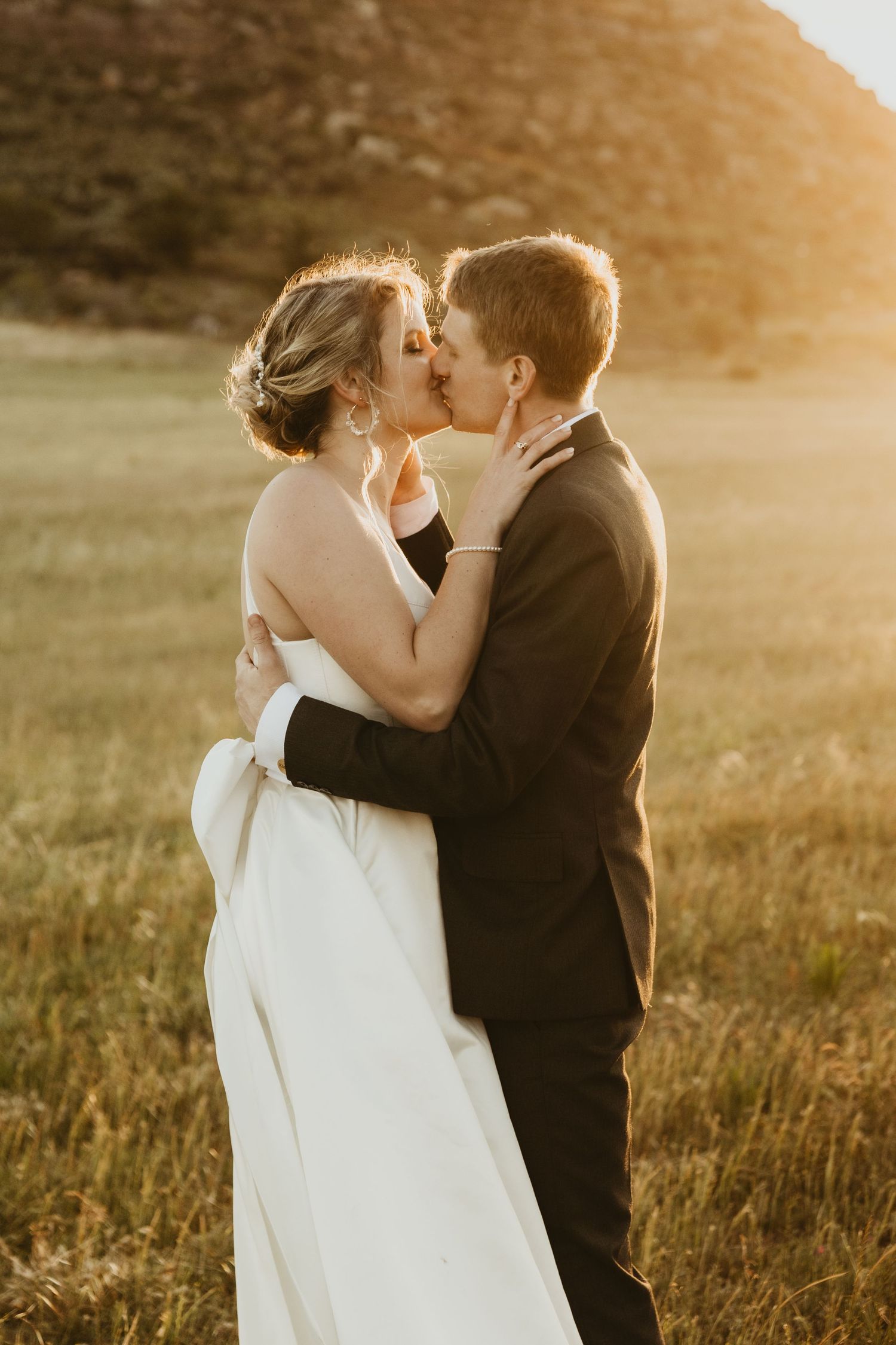 Newlywed couple embraces and kisses in golden sunlit field during sunset wedding photoshoot.