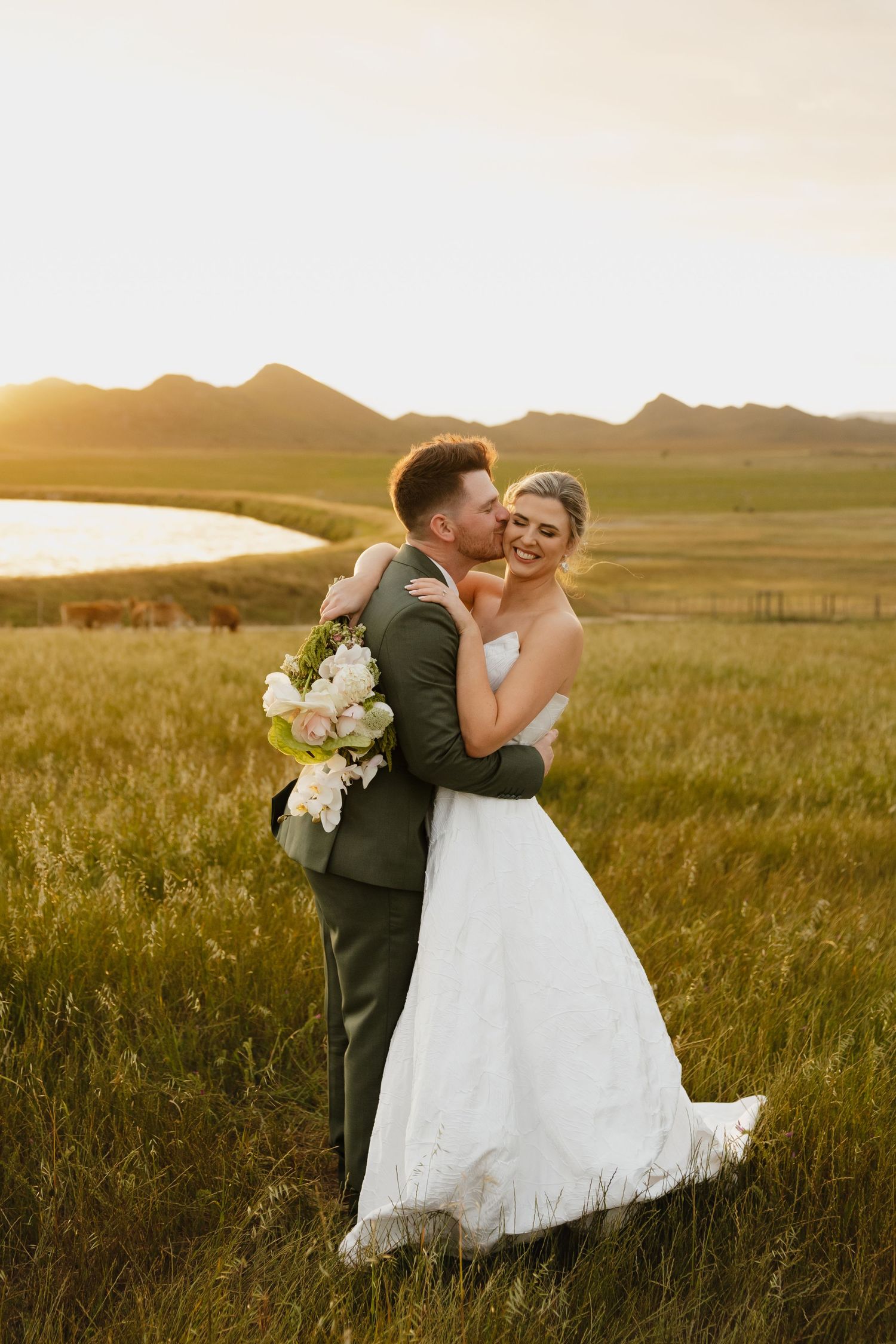 A couple in wedding attire embrace in a golden field at sunset with mountains in the background.