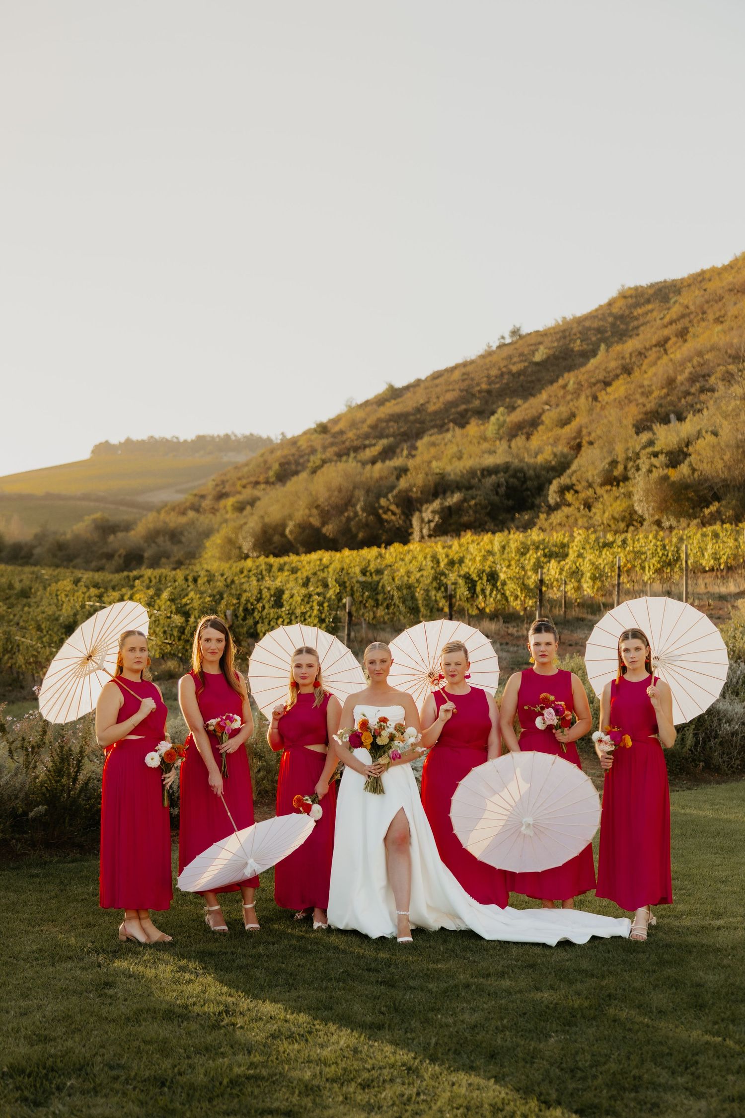Bride in white gown with bridesmaids in red dresses holding white parasols at a vineyard wedding.