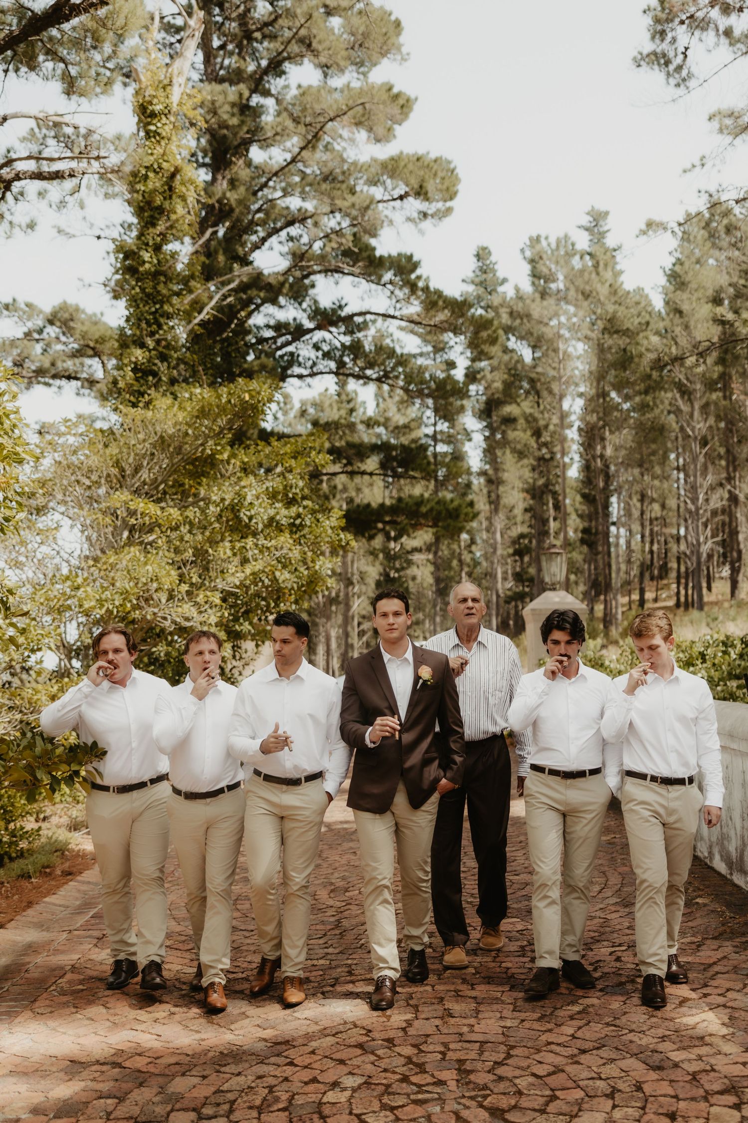 Groom in brown suit walks with groomsmen in white shirts and khaki pants on brick path surrounded by tall trees.