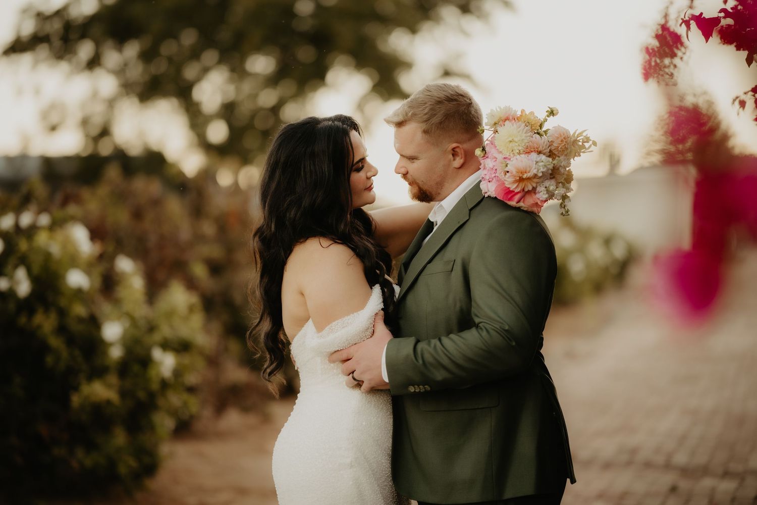 Bride and groom share a romantic moment outdoors, with bride in white gown and groom in green suit holding a floral bouquet.