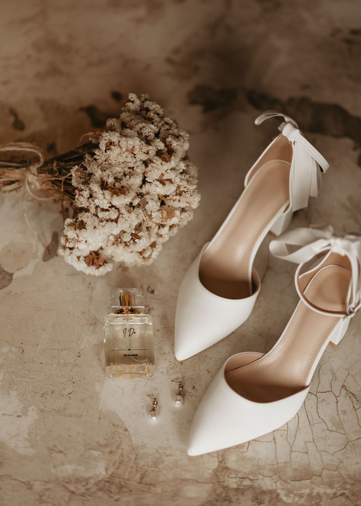 Elegant white wedding heels with bow details displayed with a perfume bottle and dried flower bouquet.