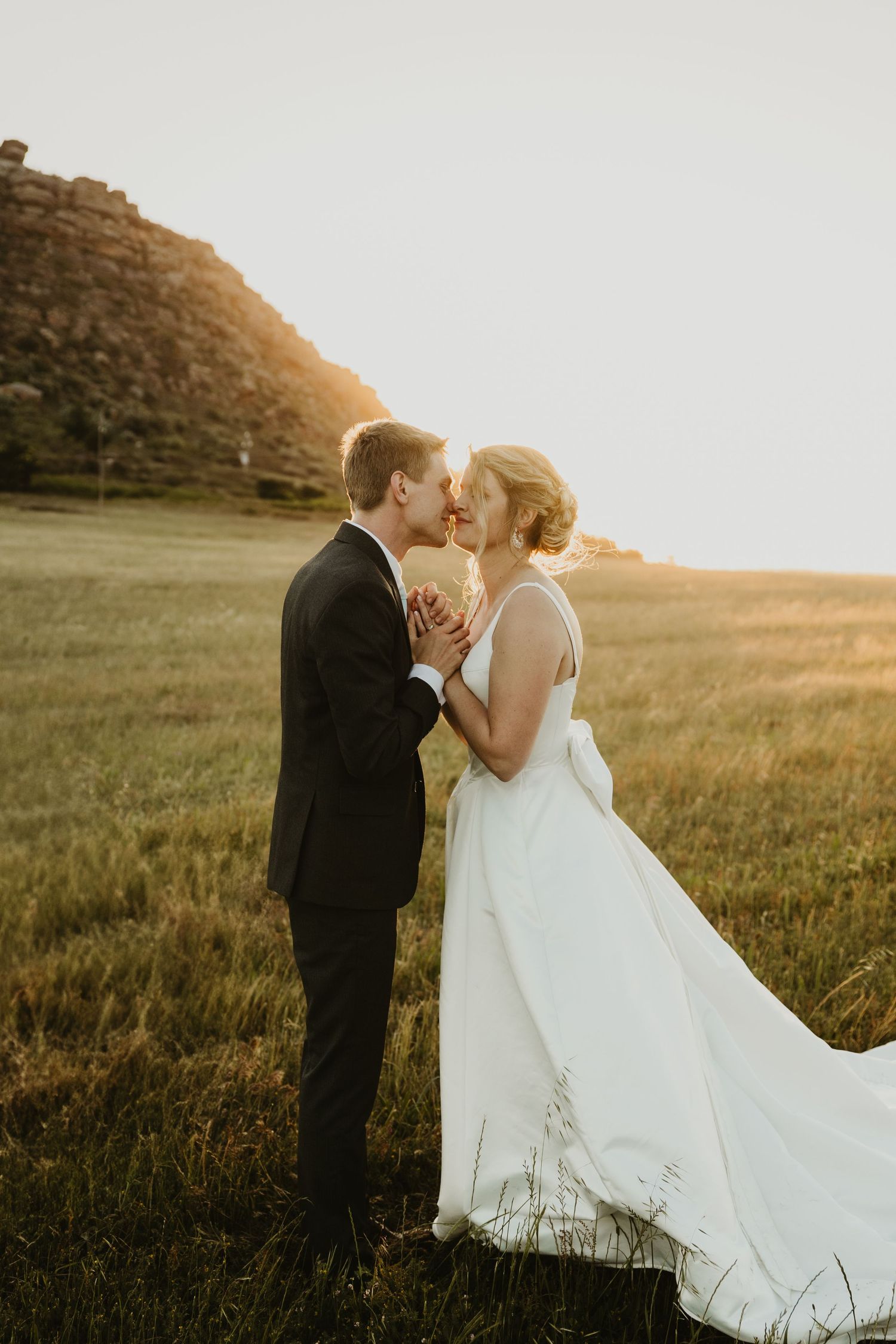 A couple embraces in a golden field at sunset while the bride's white dress flows elegantly in the breeze.